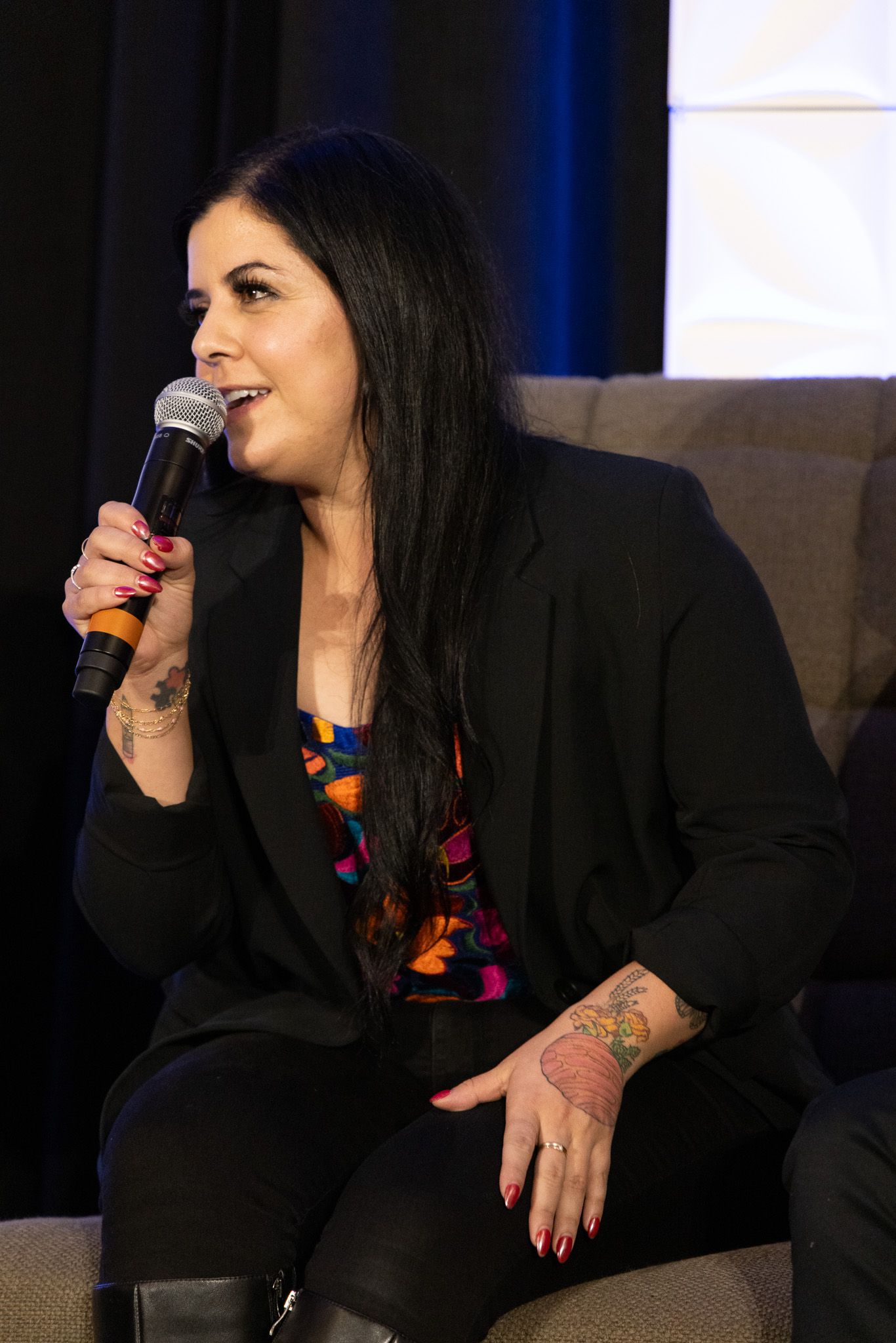 Speaker in black blazer and colorful top addresses audience using handheld microphone while seated on stage.