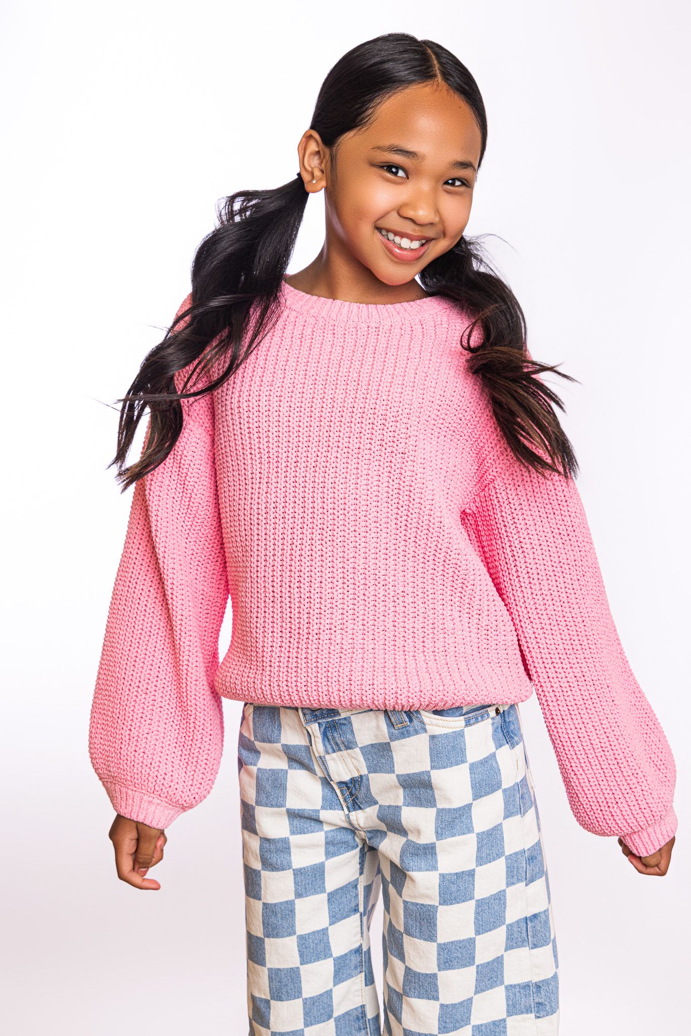 Smiling girl wearing a pink chunky knit sweater and blue checkered wide-leg jeans against a white background.