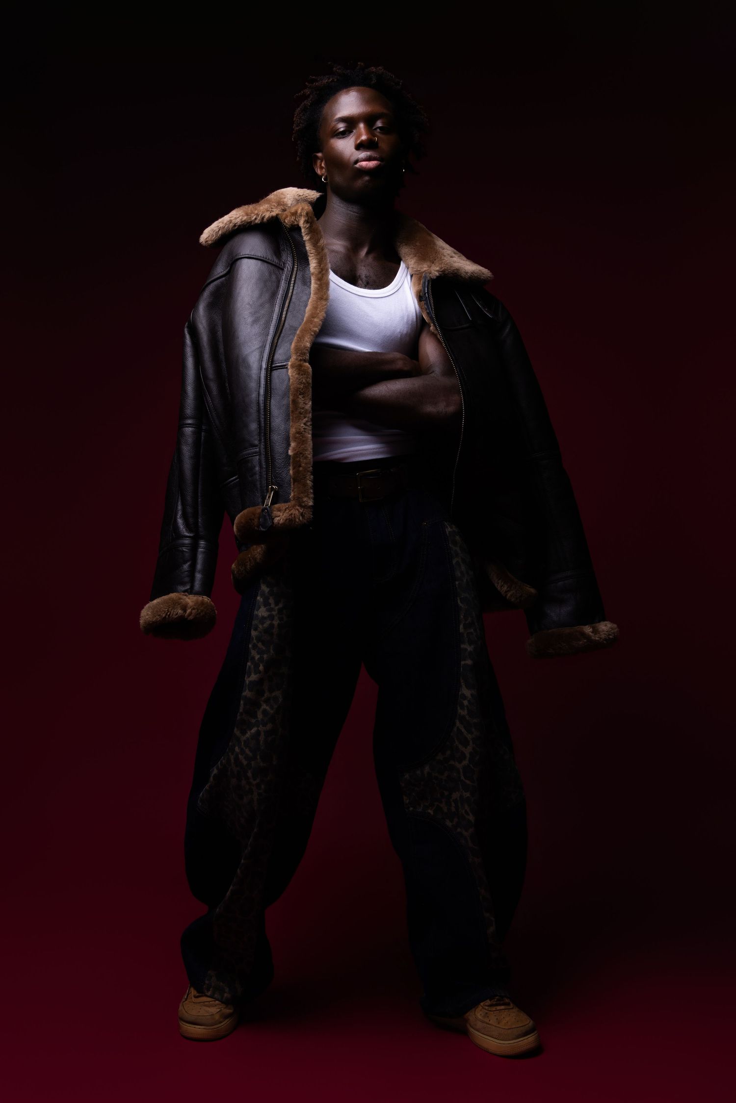 Young man posing in oversized leather shearling jacket, white tank top, and leopard print wide-leg pants on dark red background.