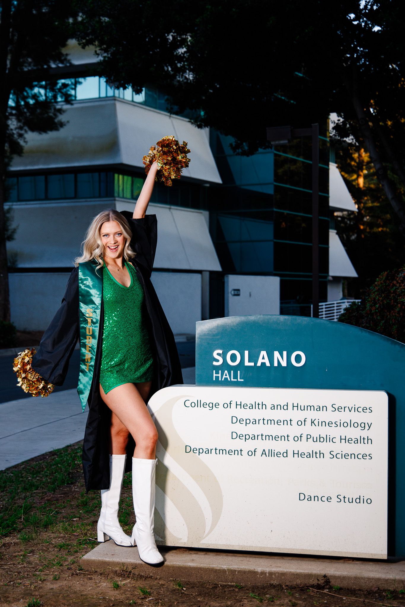 Cheerleader in green sequin dress poses with gold pom-pom by Solano Hall sign at university campus.