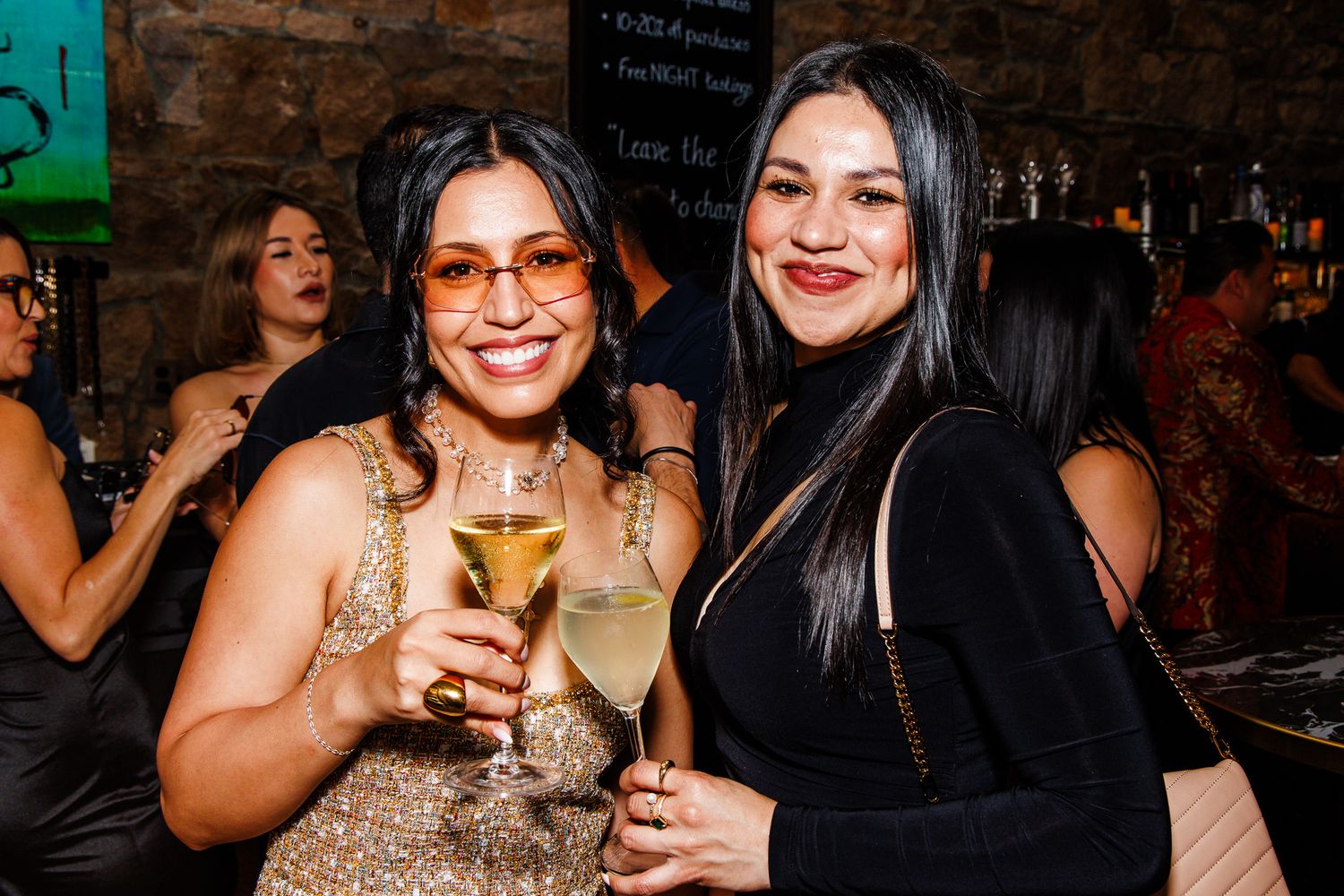 Two women toast with champagne glasses at a lively bar event, dressed in elegant party attire.