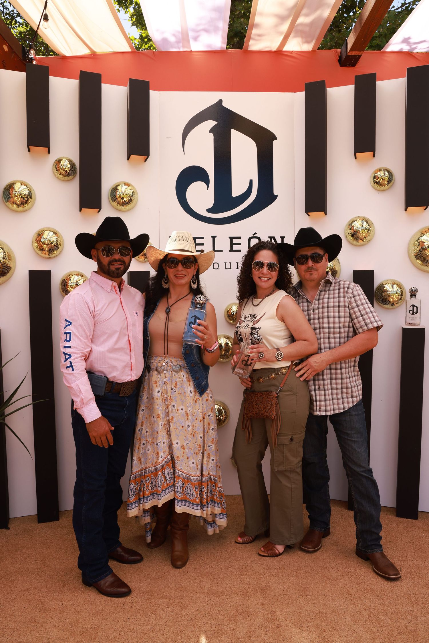 Four people in cowboy hats pose at a DeLeon Tequila branded backdrop with gold disco balls.