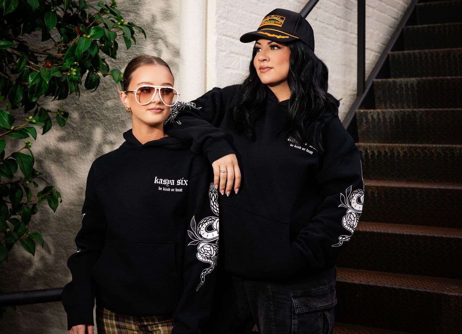 Two women wearing matching black graphic hoodies with dragon sleeve prints, posing on stairs outdoors.