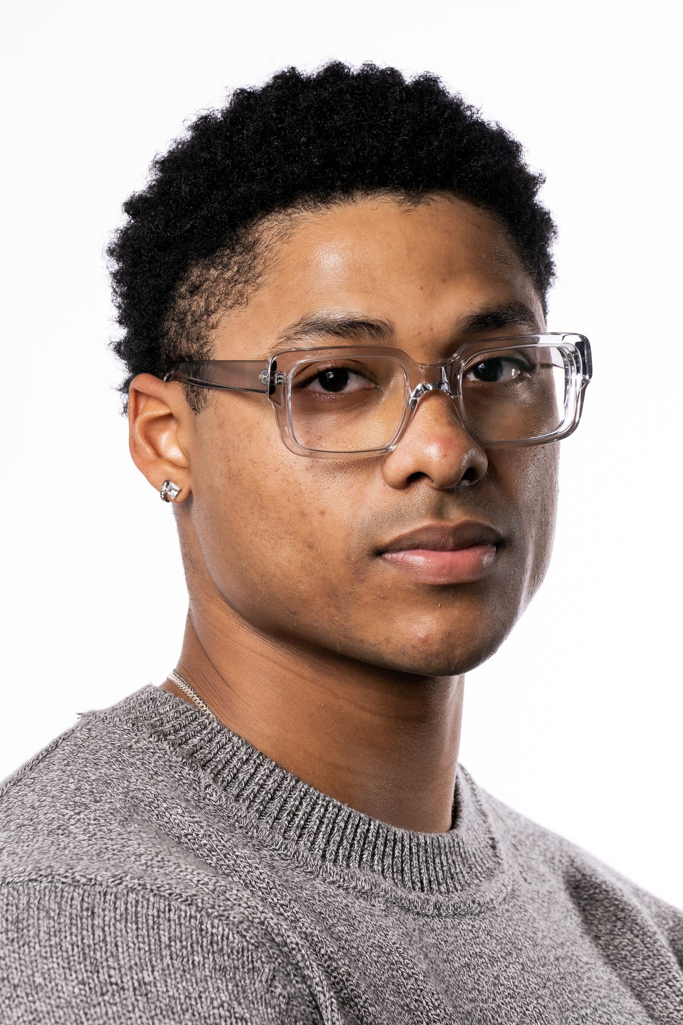 Young Black man with afro hair wearing clear-frame glasses and gray sweater poses against white background.