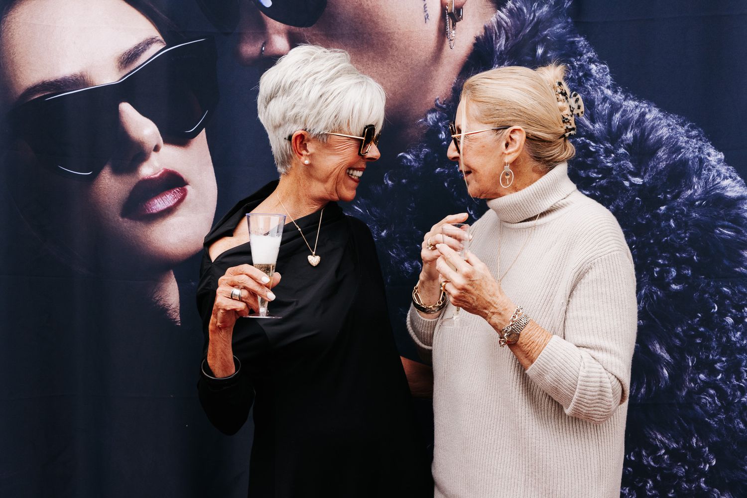 Two elegant older women laugh together at an event, one holding a champagne glass, standing before a fashion backdrop.