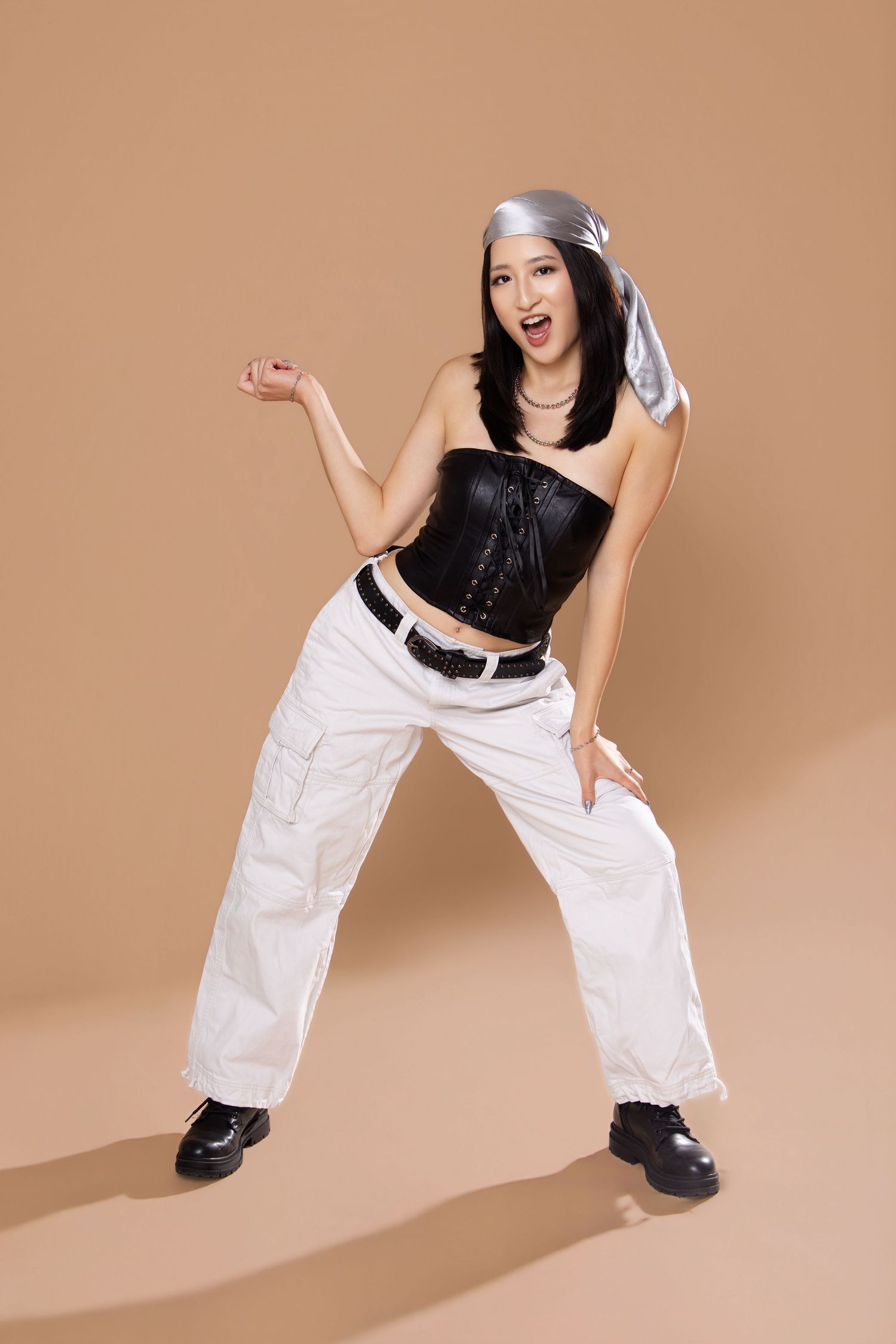 Young woman in black corset top, white cargo pants, and black boots poses dynamically against a beige background.