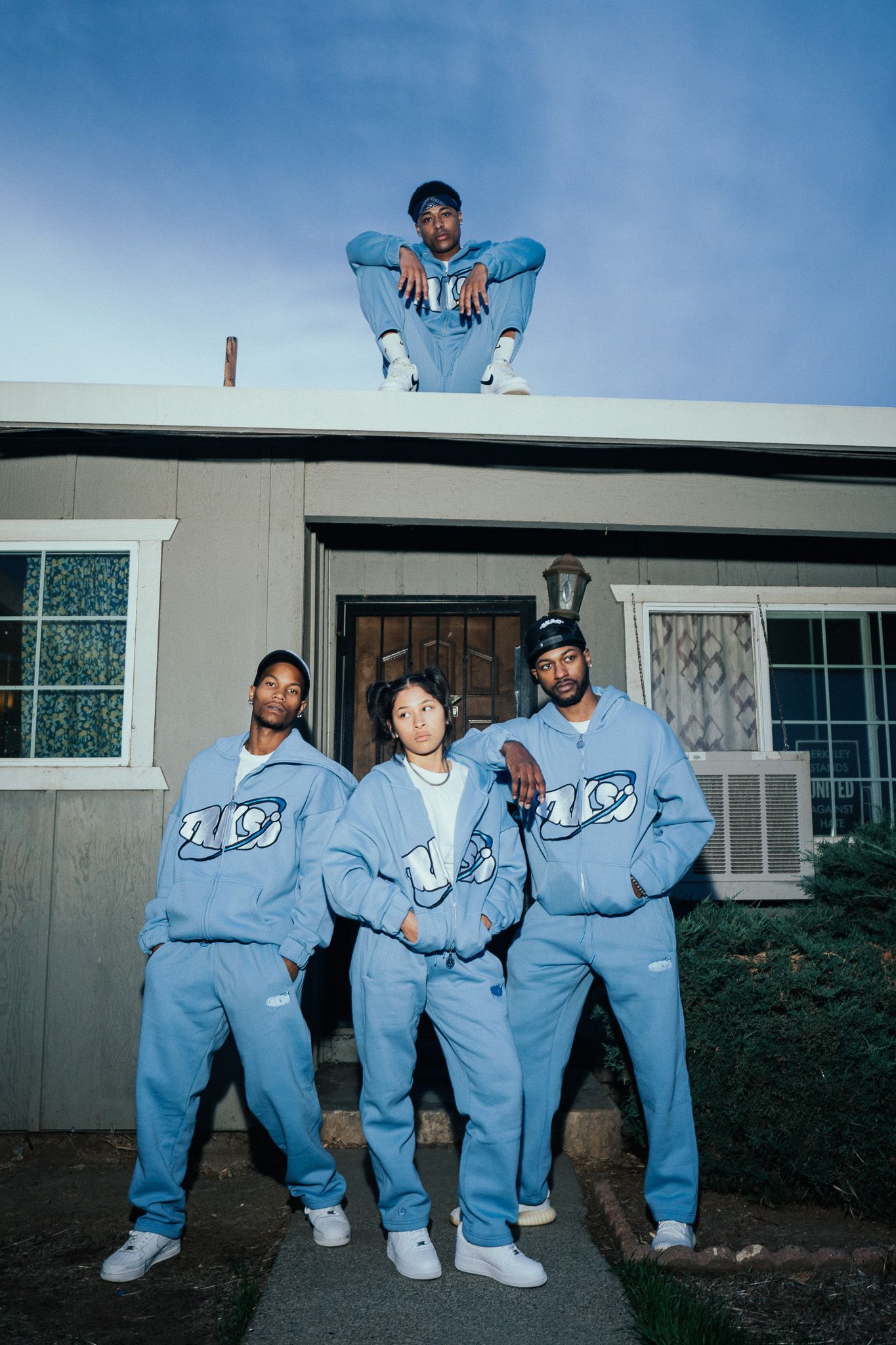 Four people in matching blue tracksuits pose outside a house, one sitting on the roof.