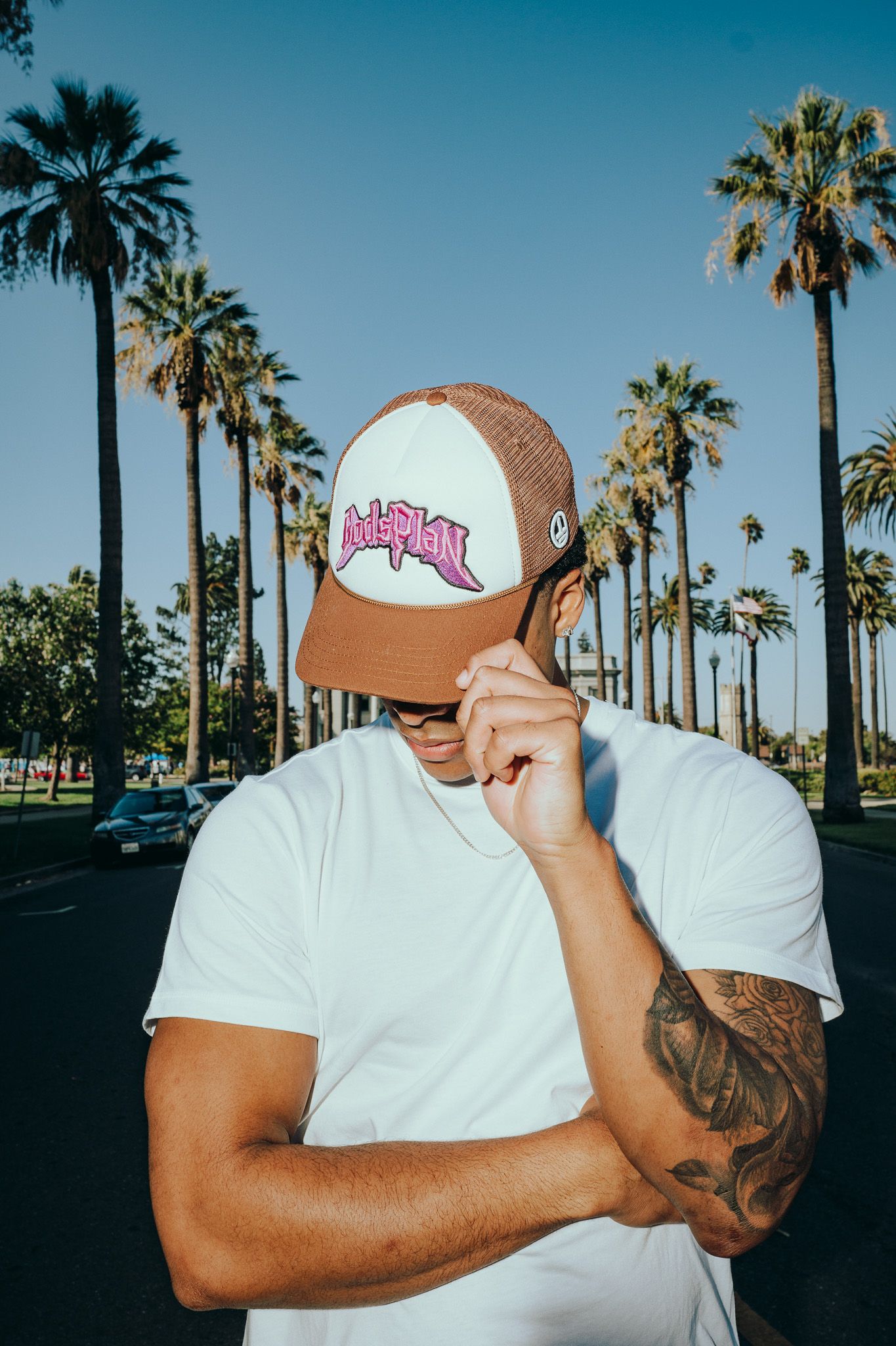 Man in white tee and brown trucker hat with pink logo poses among palm trees, showcasing arm tattoos.