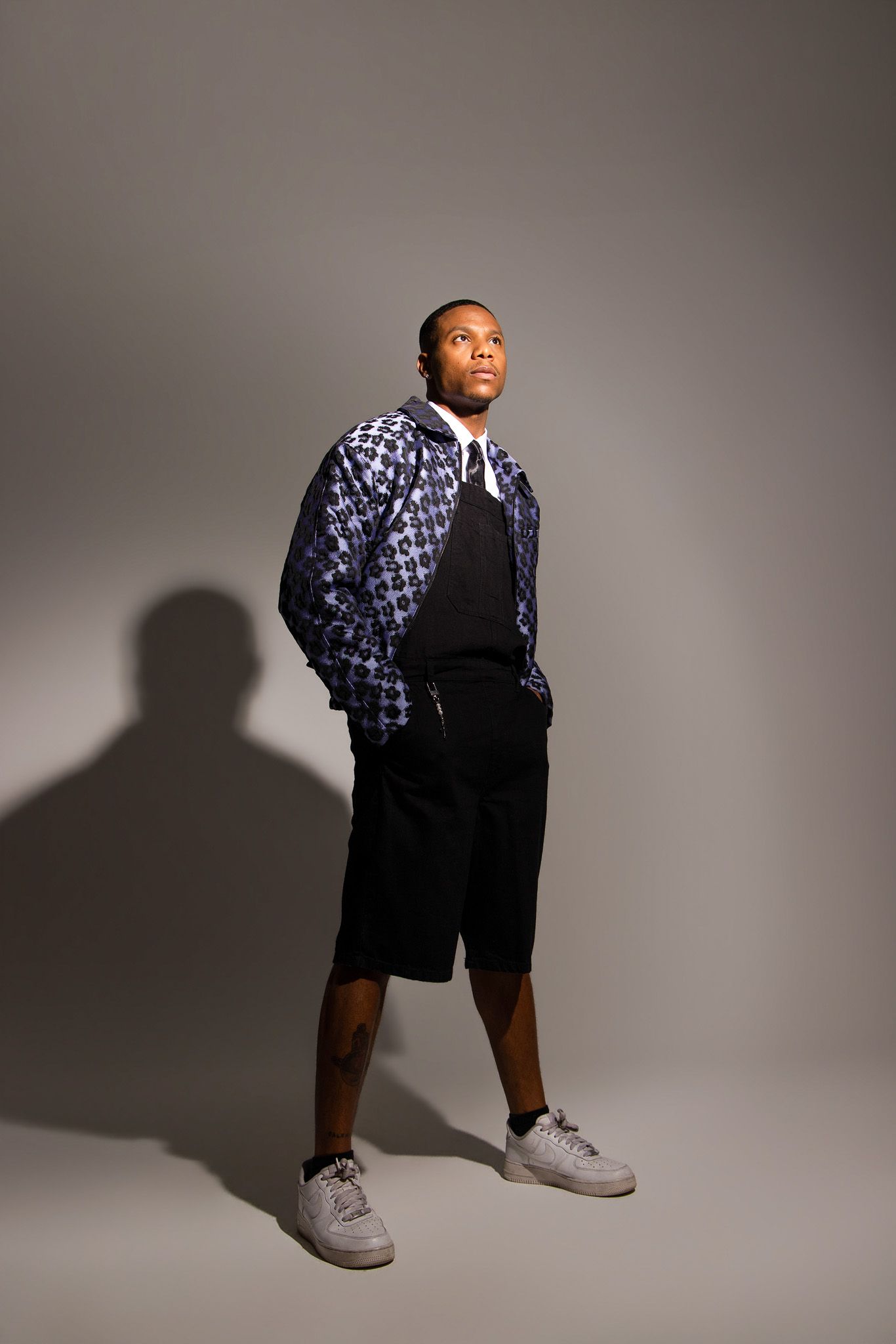 Studio fashion photo of someone wearing a patterned jacket, black shorts and white sneakers with dramatic shadow on gray backdrop.