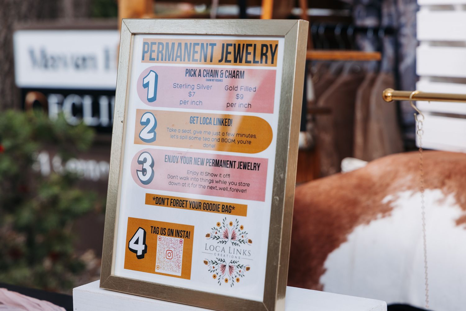 A framed permanent jewelry sign displaying four steps for pick a chain, clasp, engrave, and scan QR code at a boutique event.