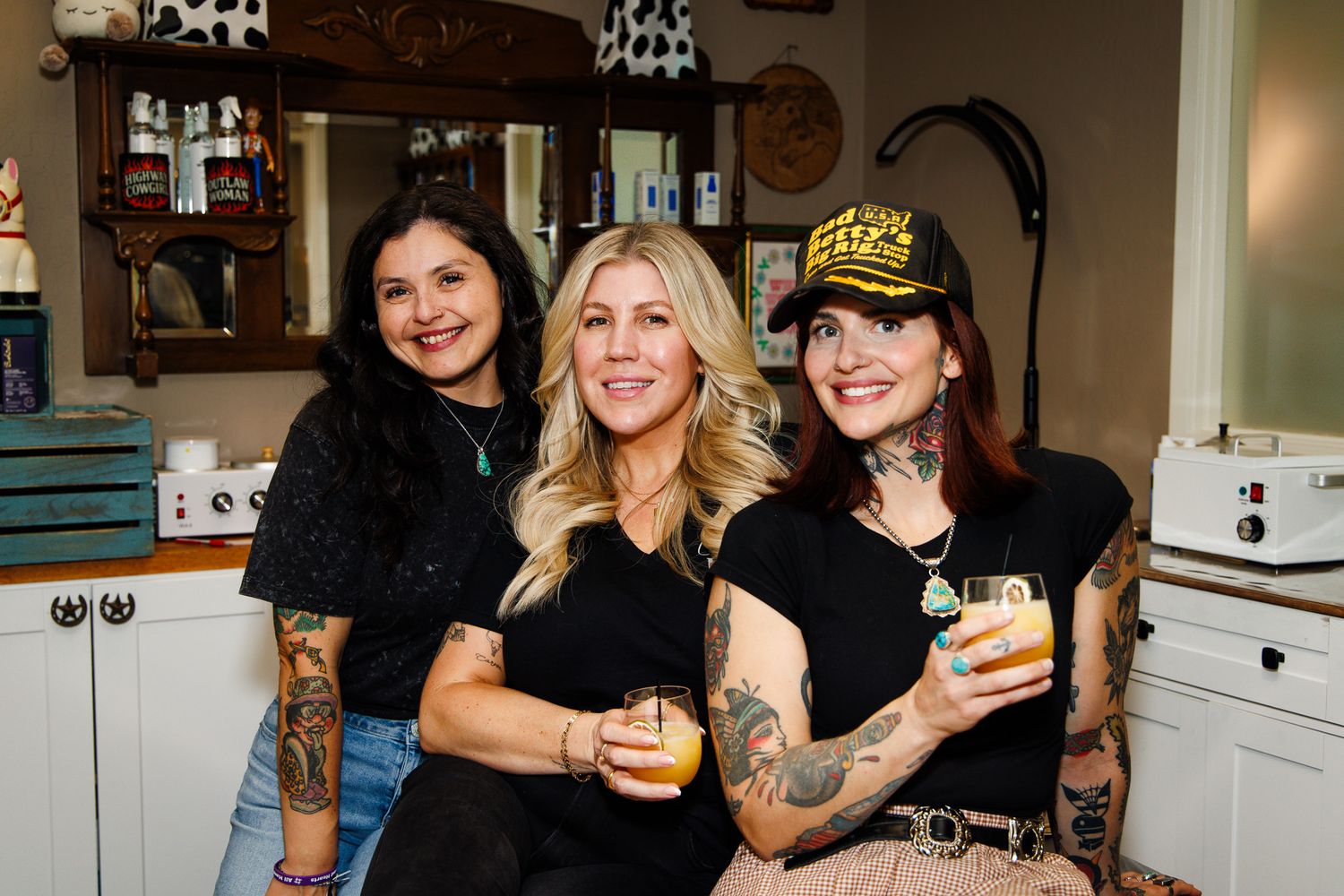 Three tattooed women smiling and holding drinks together inside a cozy decorated salon space.