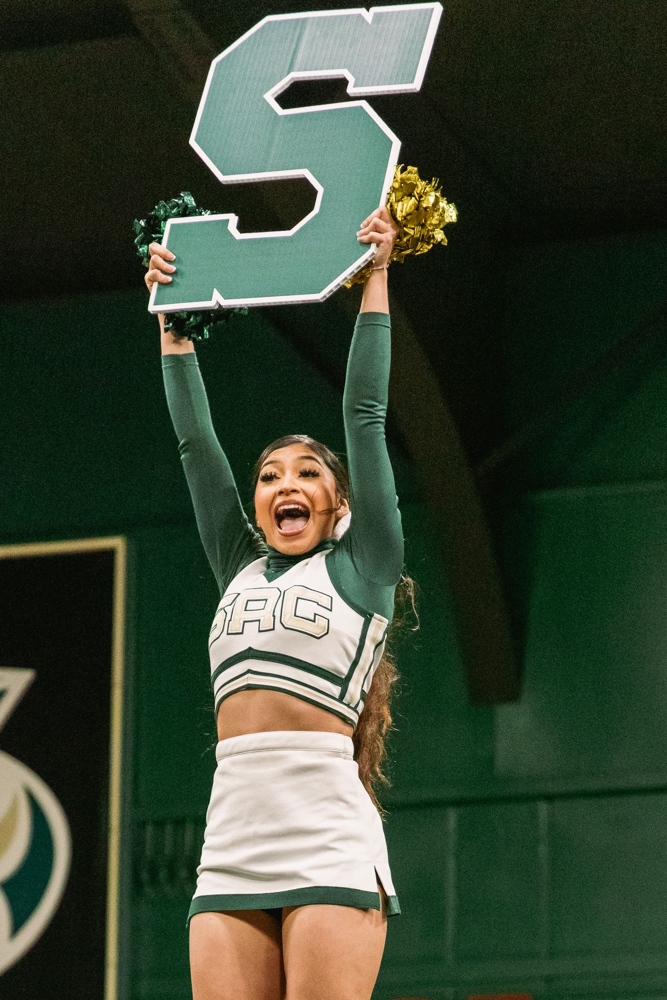 Sacramento State cheerleader holds letter S and gold pom poms in green uniform.