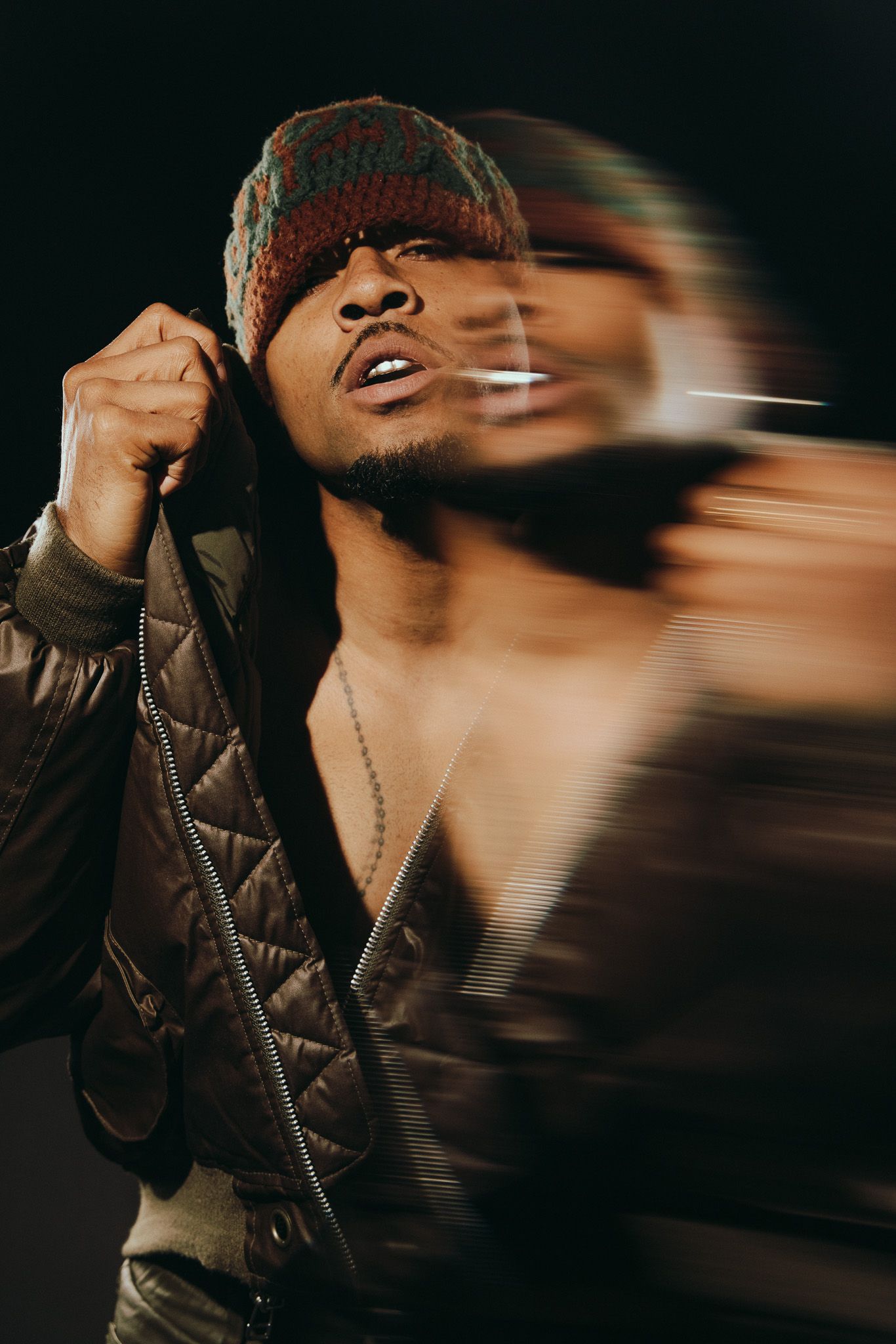 Young Black man in quilted jacket and colorful knit hat poses dramatically with motion blur effect on dark background.