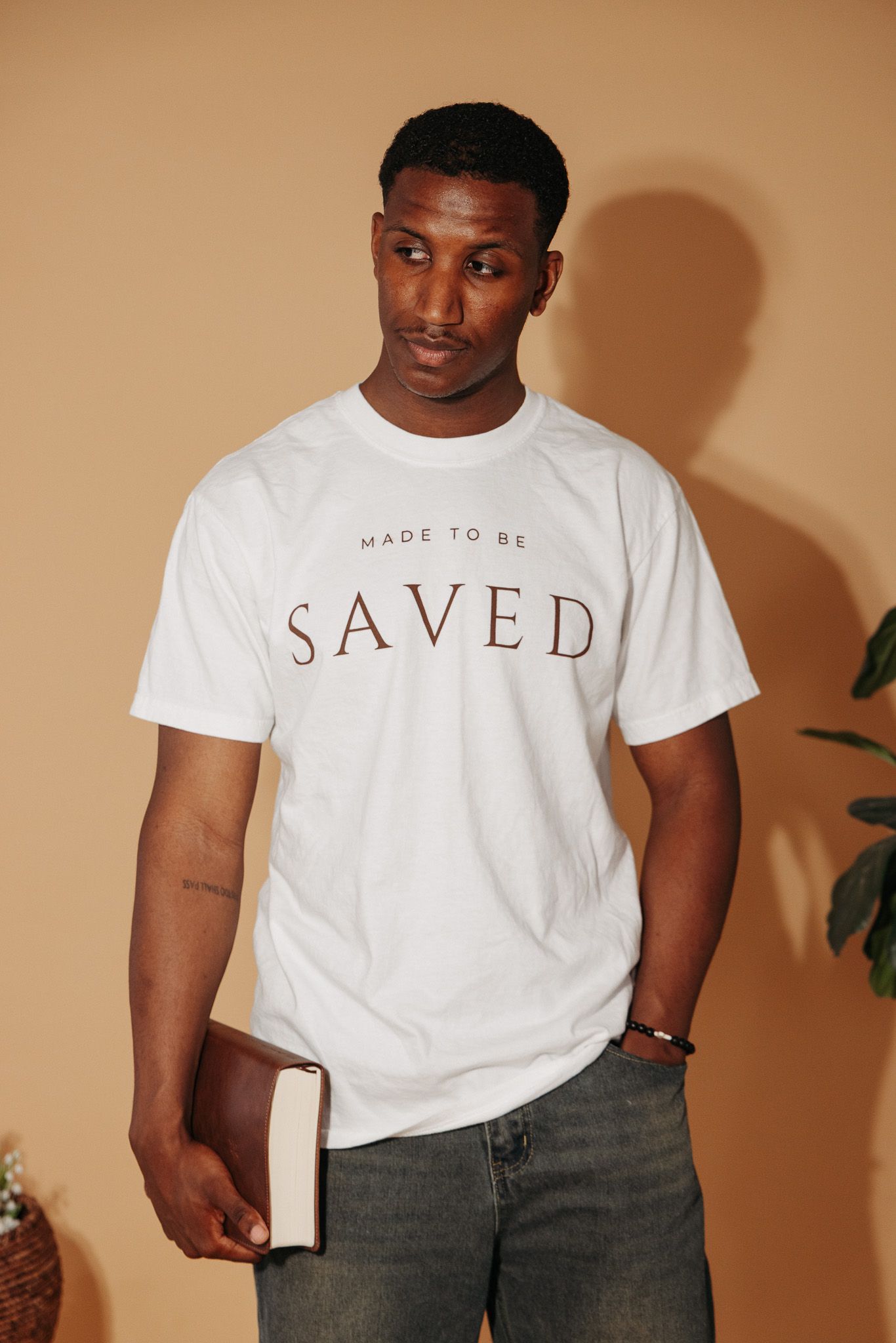 A white t-shirt collection featuring minimalist text designs 'Made To Be' and 'Saved' against a warm beige background.