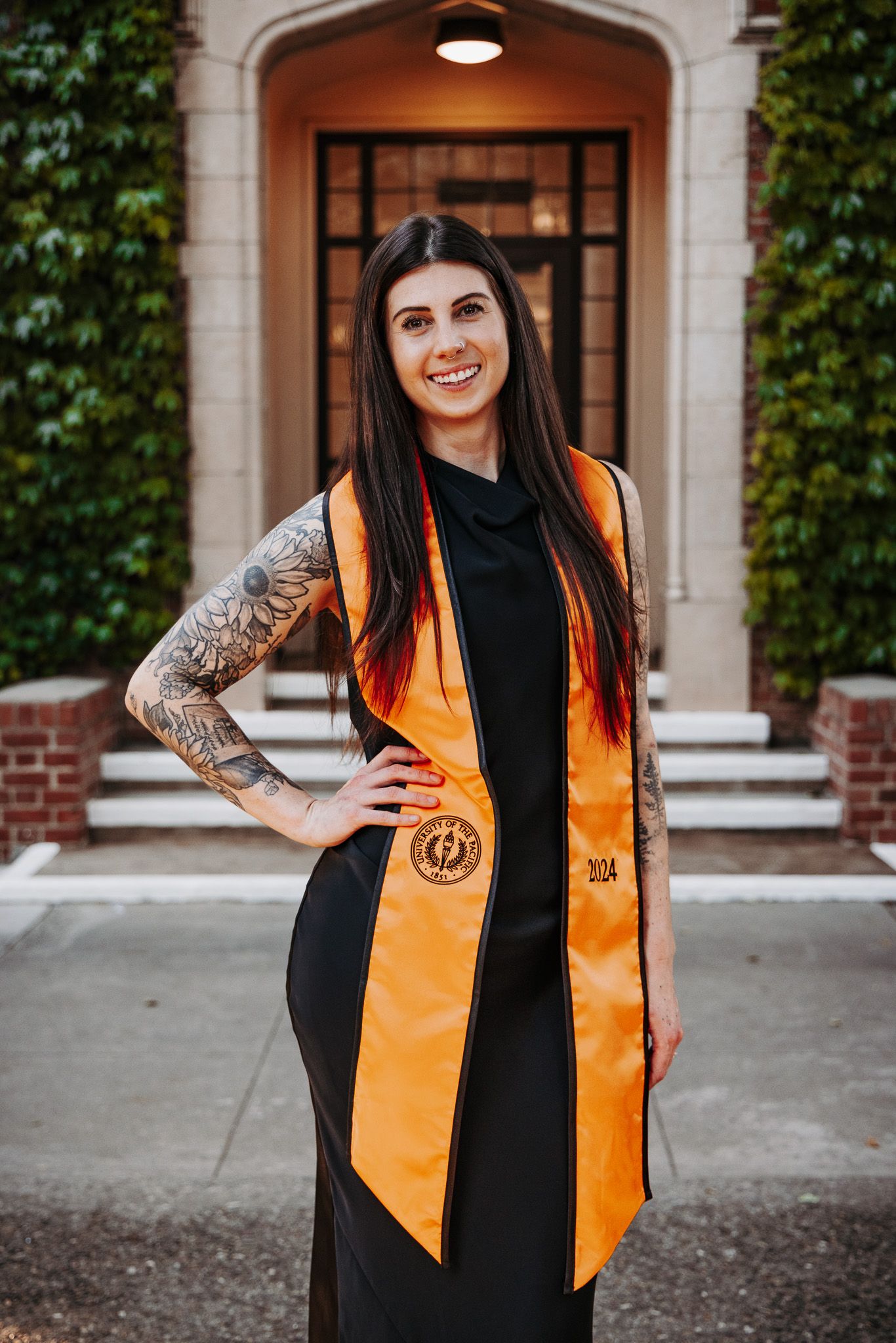 A graduate wearing a black dress and orange graduation stole poses for photos against a brick building with ivy.