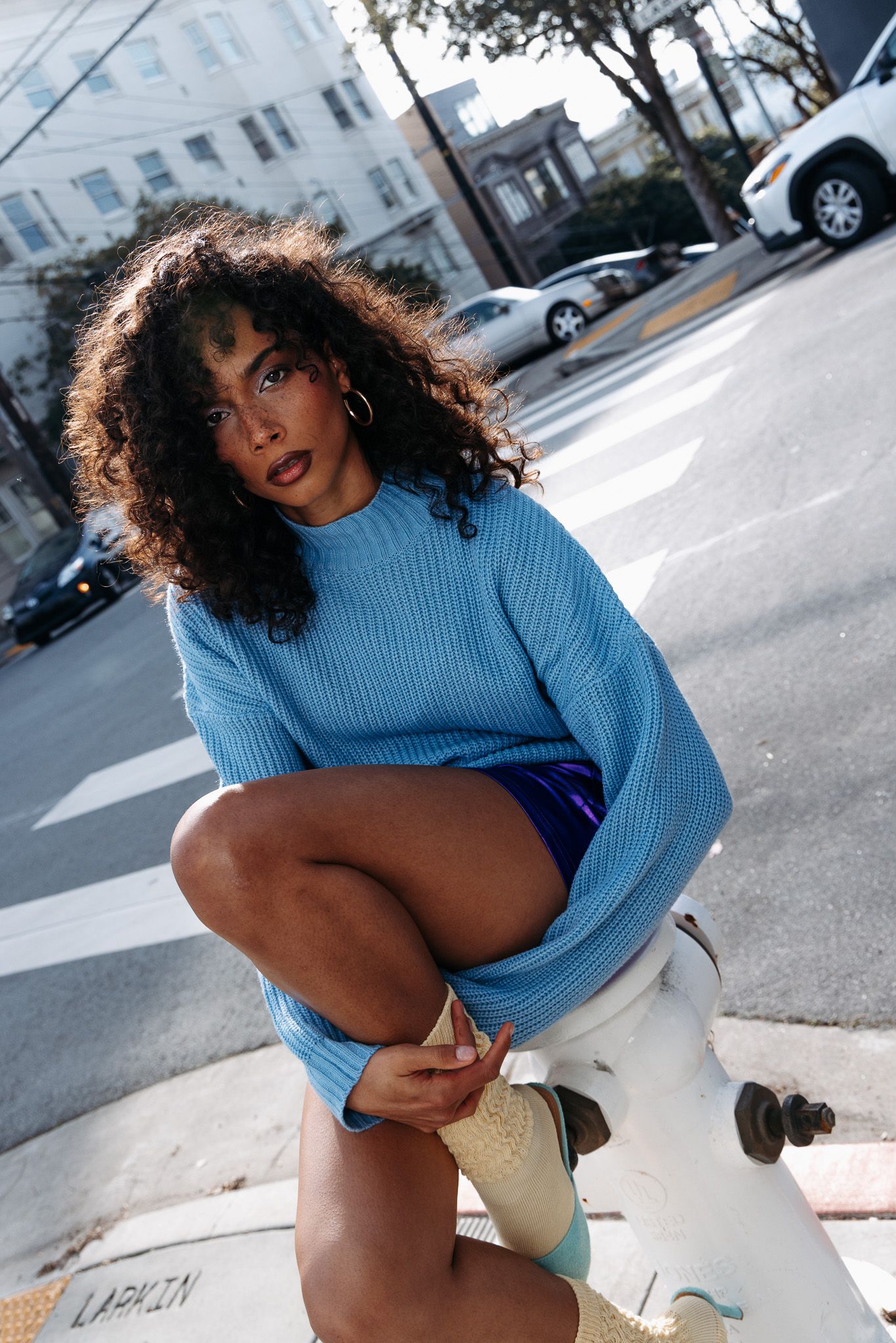Street style photoshoot featuring blue knit sweater and white boots against city backdrop.