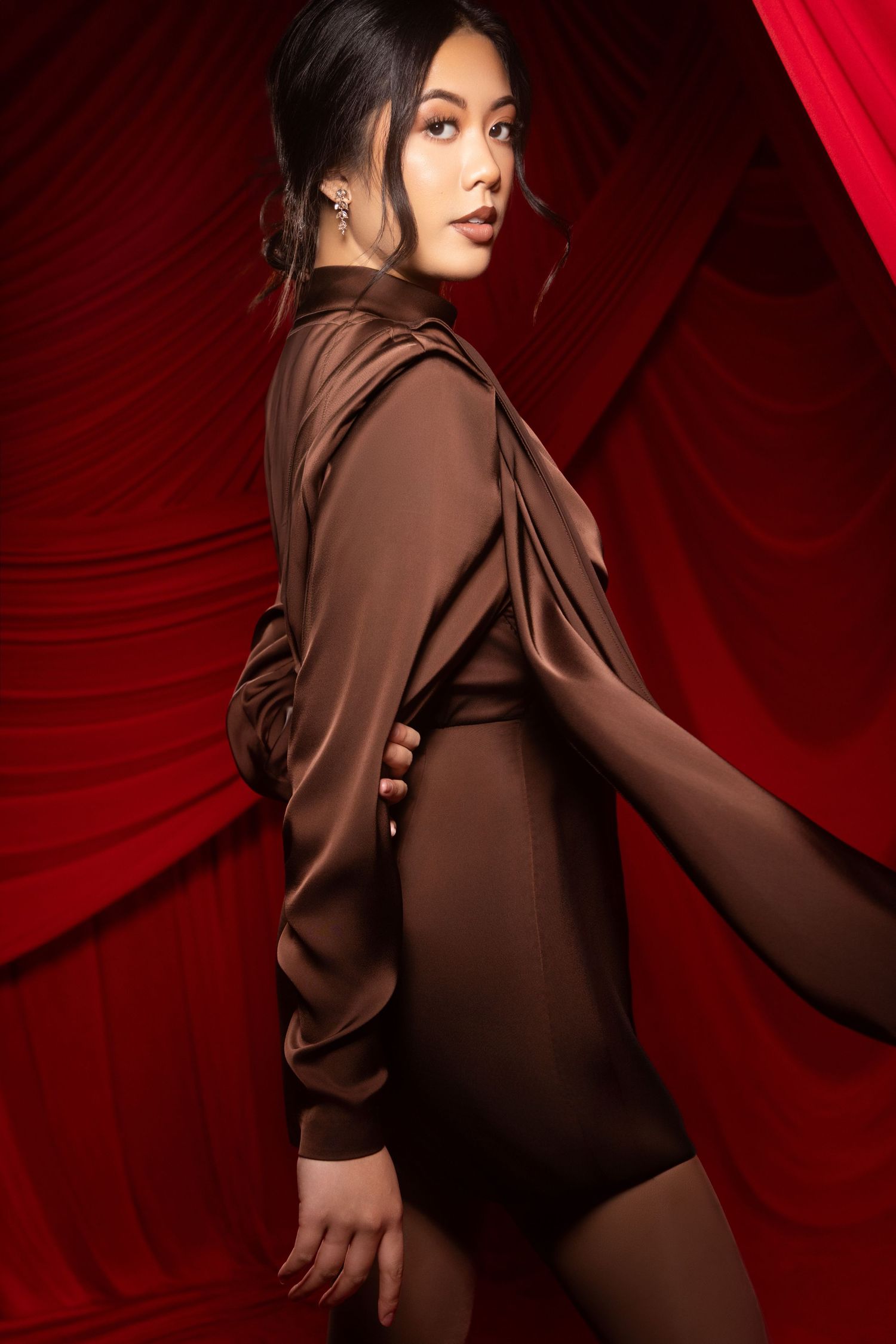 Fashion portrait of model in brown draped dress posing dramatically against red velvet backdrop.