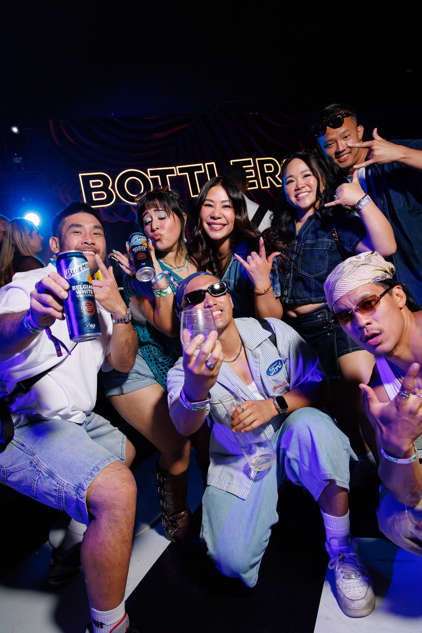 Excited group of friends posing with drinks at BottleRock festival under purple lights in dark venue.