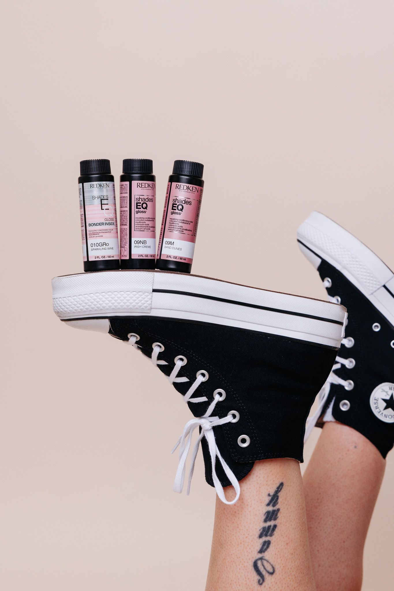 Redken hair products balanced on black Converse sneakers against a beige background, with a tattooed leg visible.