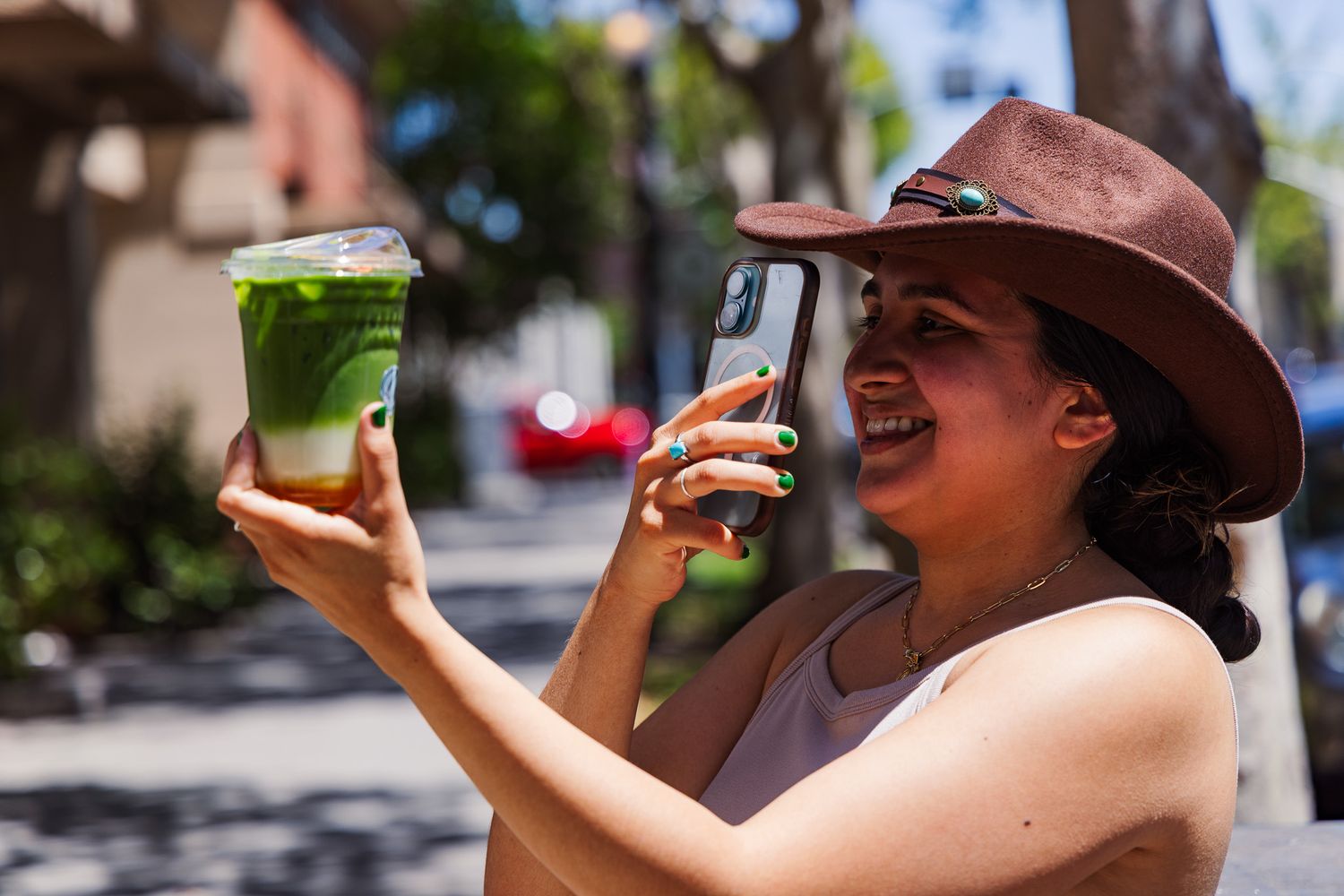 A woman in a brown hat smiles while photographing a green iced drink outdoors on a sunny day.