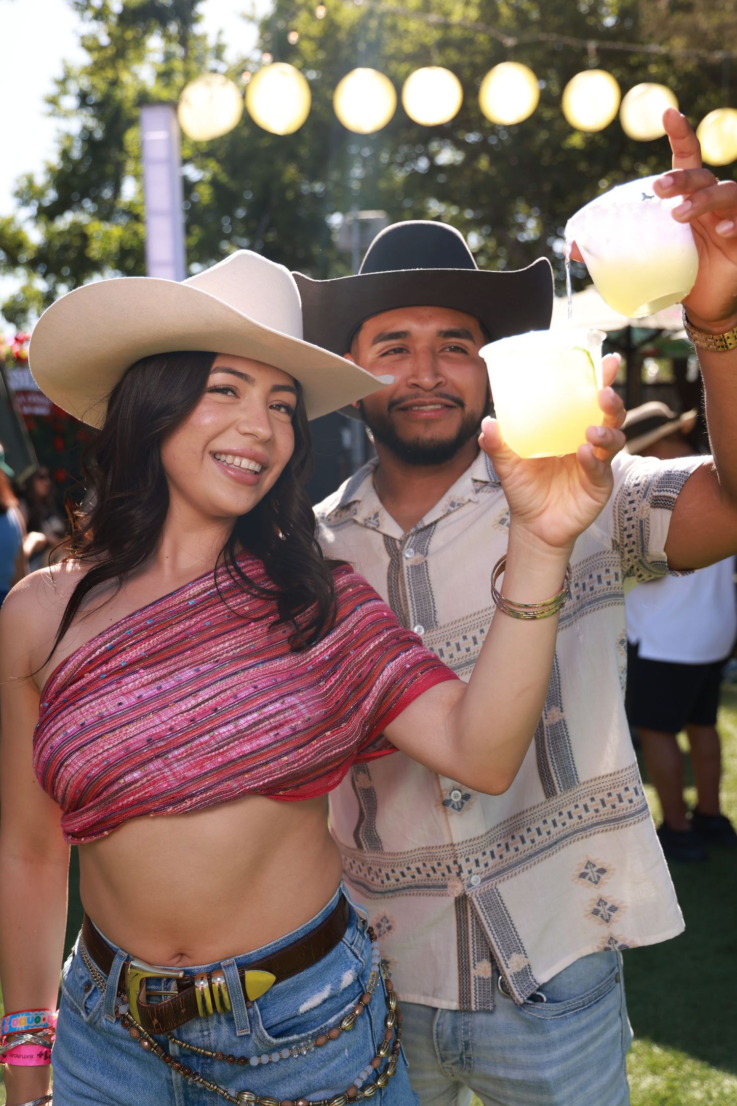 Two friends in cowboy hats smile at a festival, one raising a large frozen drink outdoors.