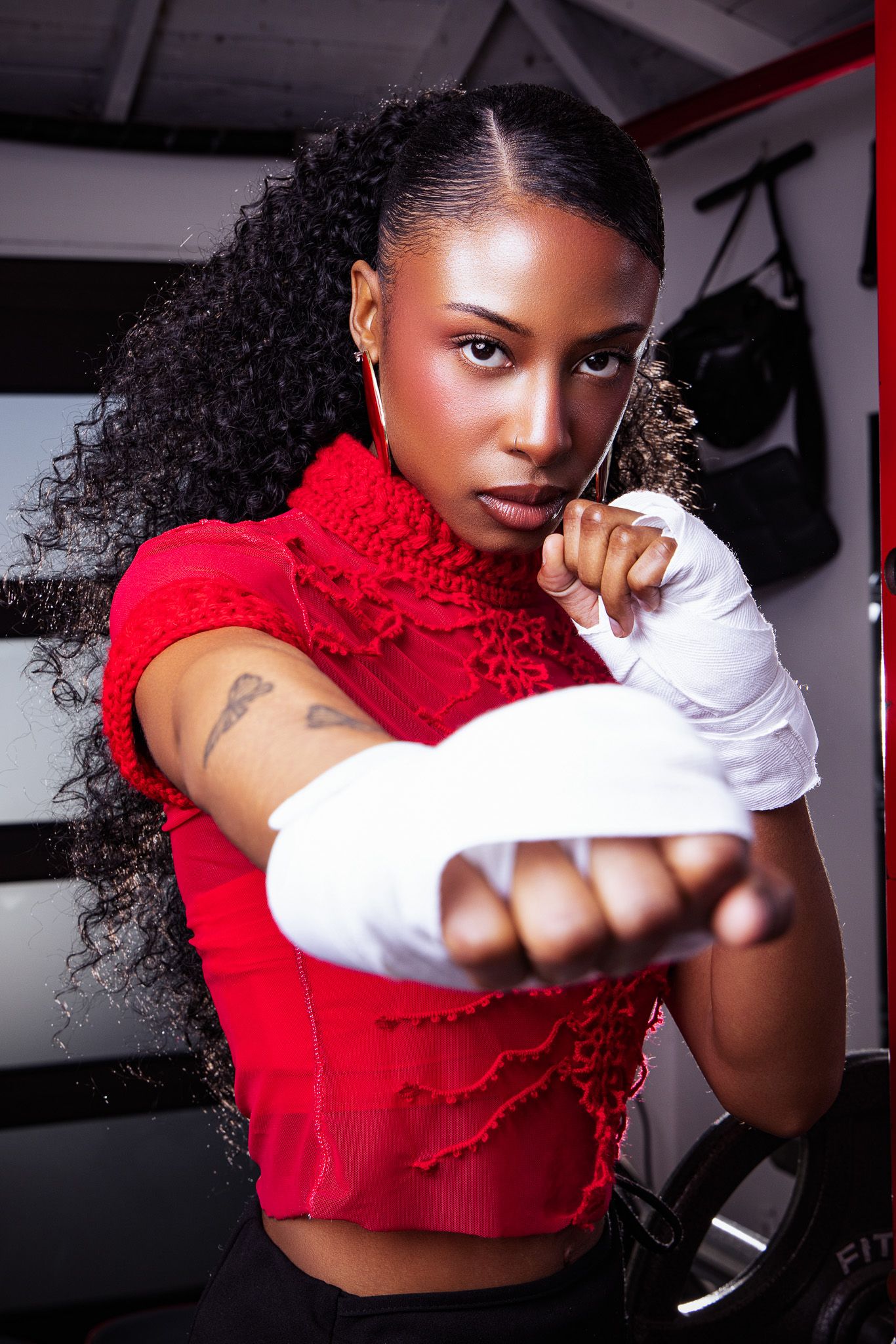 A fierce female boxer in a red top with white hand wraps throws a punch in a gym setting.