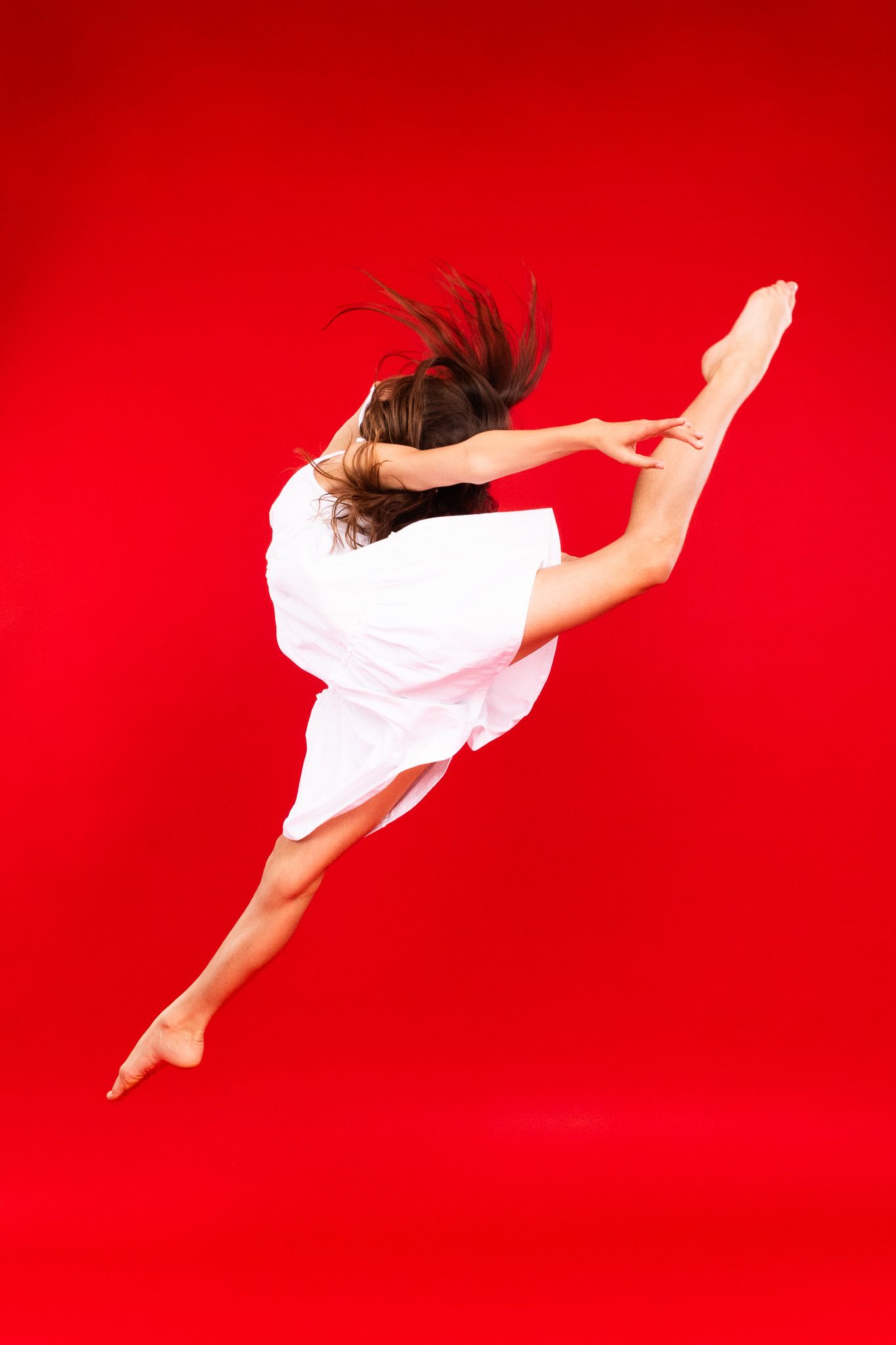 Dancer in white dress leaps gracefully through the air against vibrant red background.