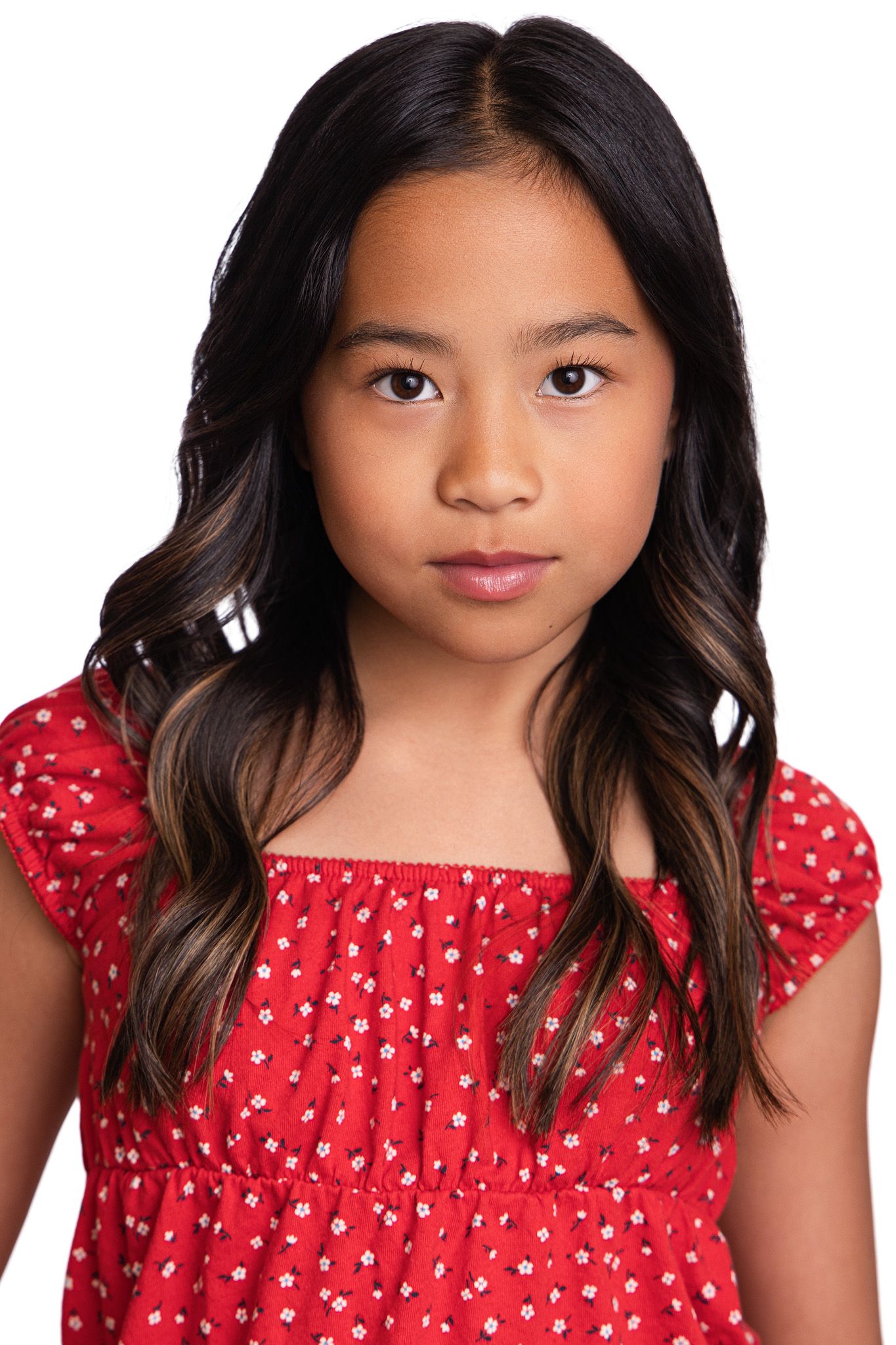 Young girl with long dark hair wearing a red floral dress poses for a portrait photo against white background.