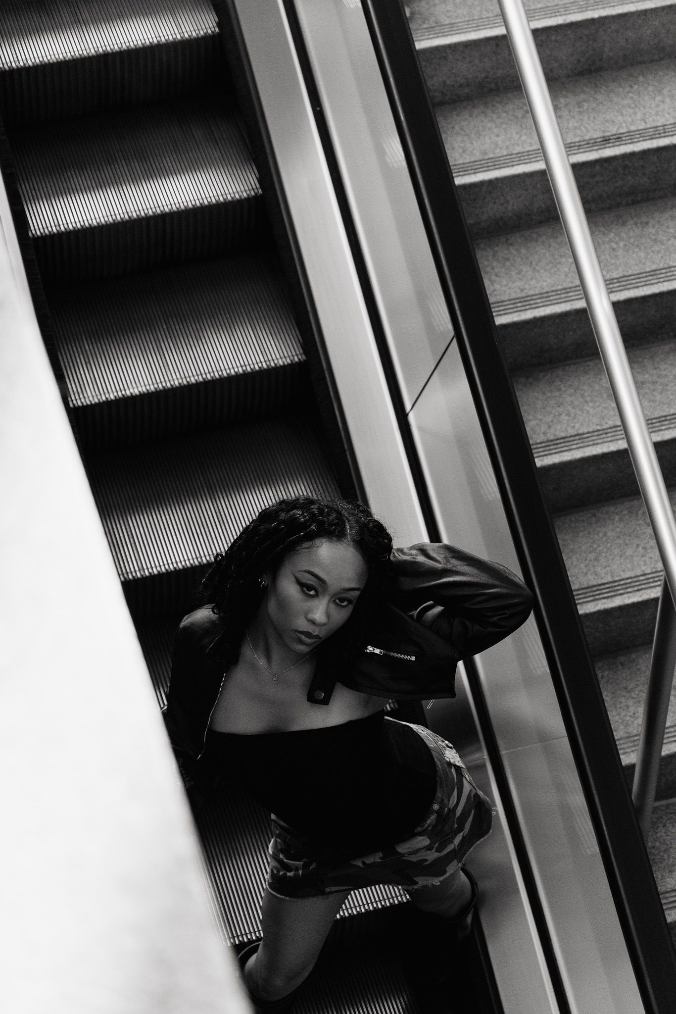 A black and white artistic portrait of a person posing on an escalator in dramatic lighting.