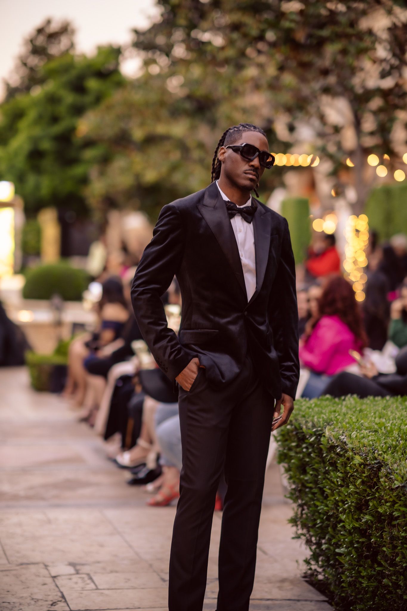 A person in a black suit stands near manicured hedges at an outdoor evening event with soft lighting in the background.