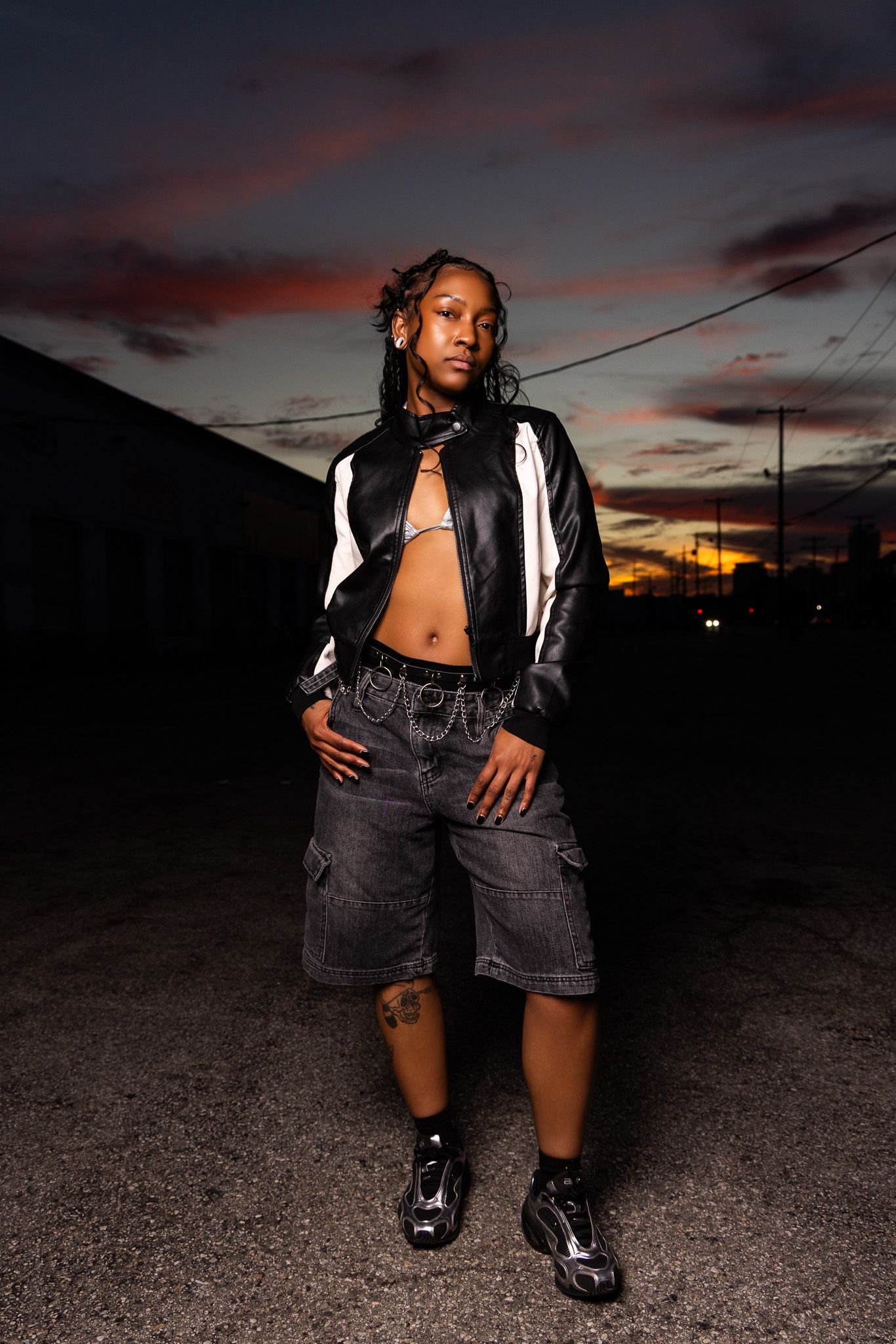 Stylish portrait at sunset showing a black and white jacket, denim shorts and shiny shoes against dramatic orange sky.