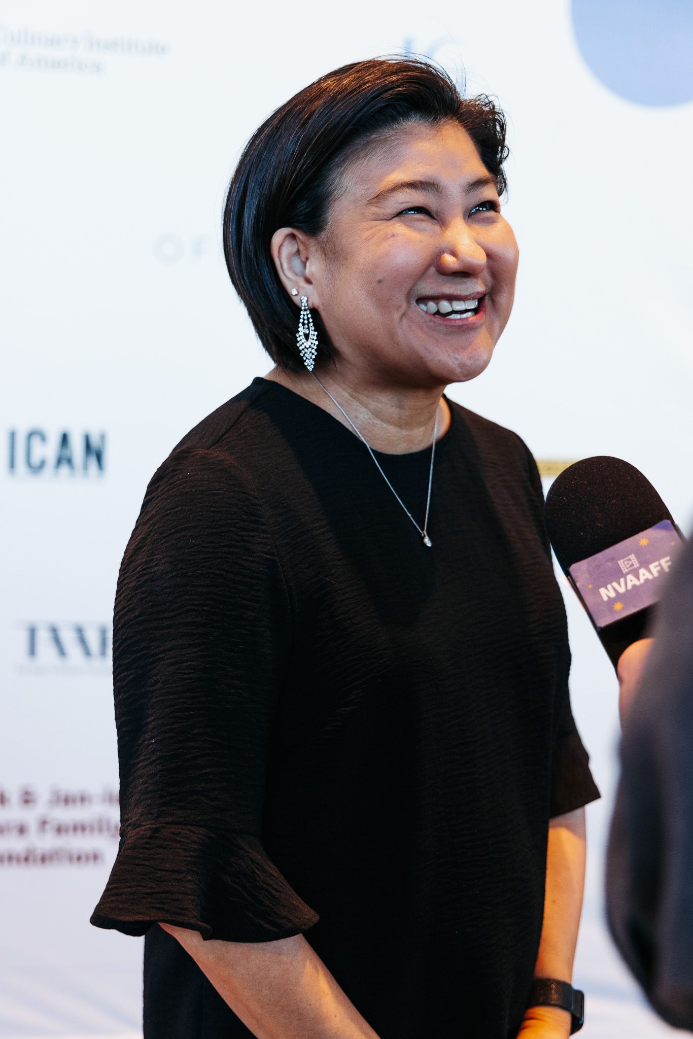 Person in black attire smiling warmly against a white backdrop at a professional event.