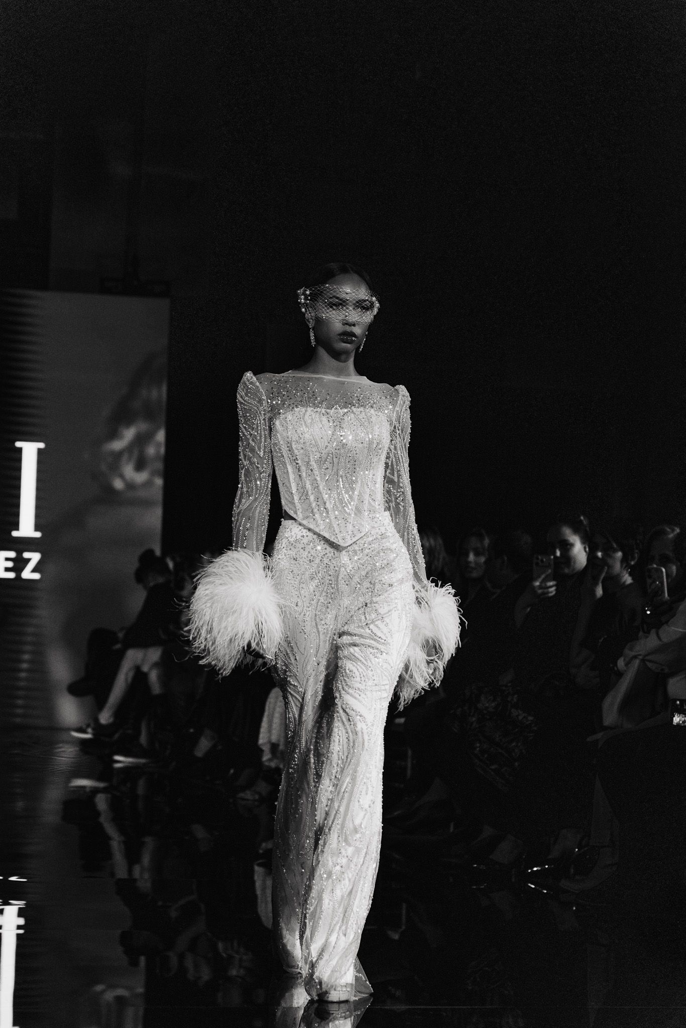Model walks runway in elegant white feathered evening gown during high fashion show.