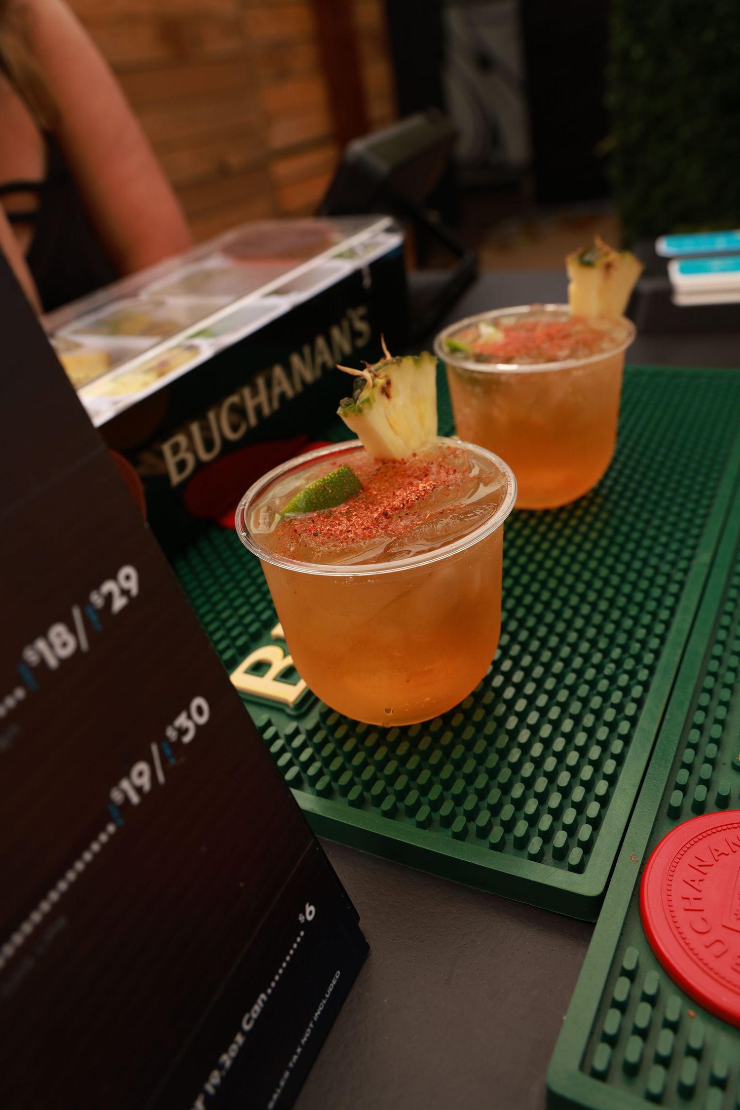 Two craft cocktails garnished with pineapple sit on a green bar mat next to a Buchanan's menu in dim lighting.