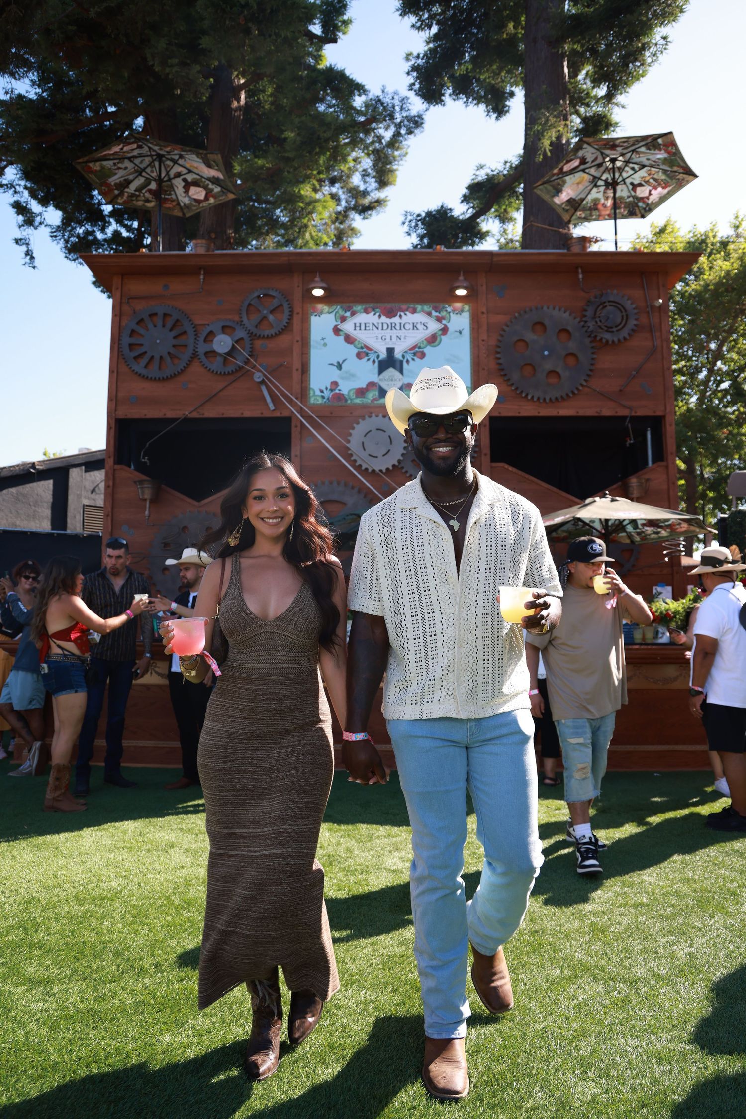A couple enjoying a sunny outdoor event with rustic wooden structures and umbrellas in the background.