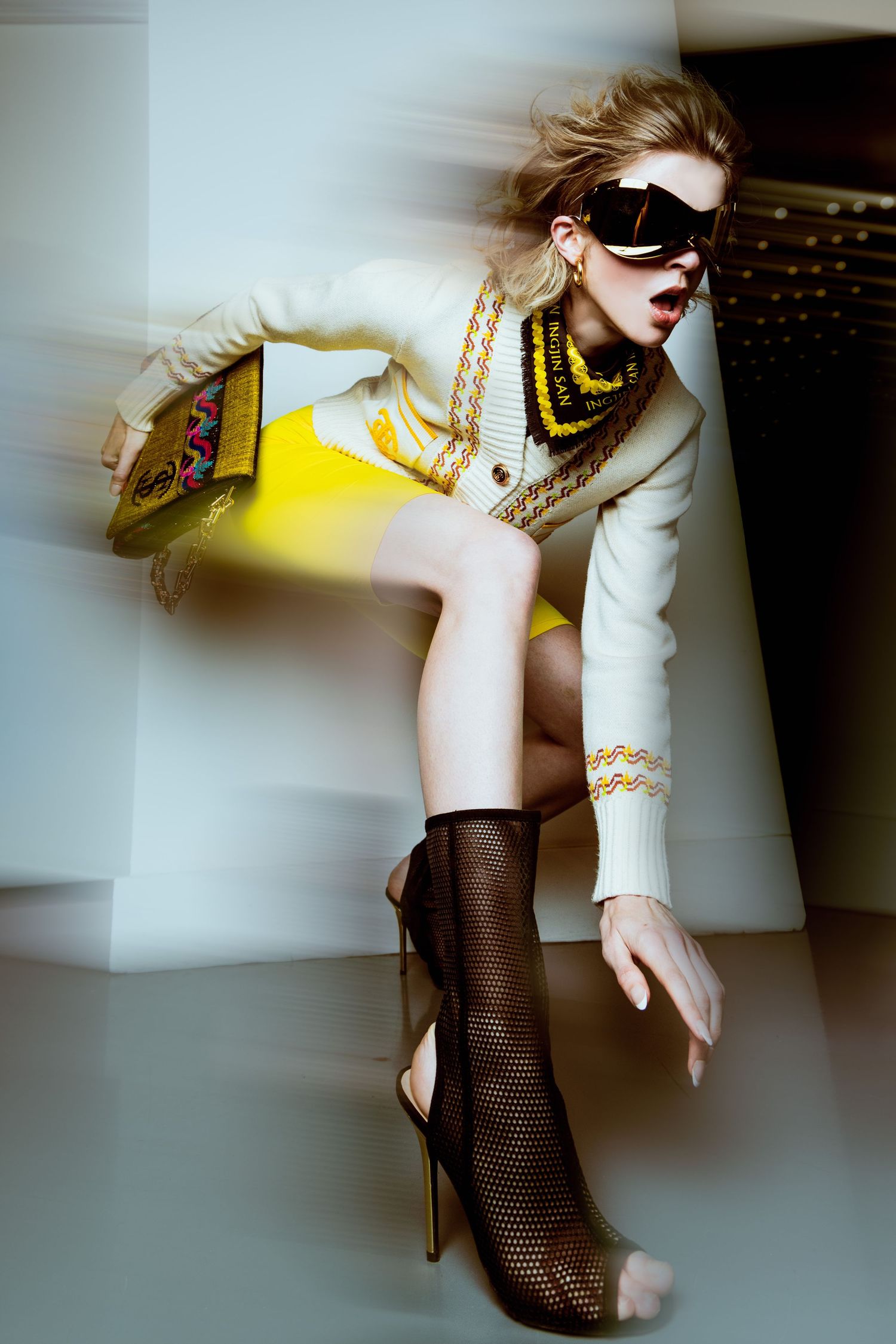 Fashion model in white jacket and mesh stockings poses dynamically against light background in editorial style shoot.