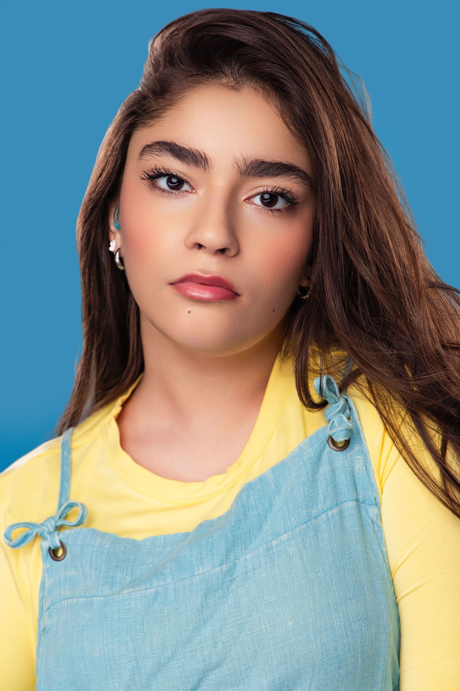 Portrait against blue background wearing yellow collar and light blue top with flowing dark hair and red lipstick.