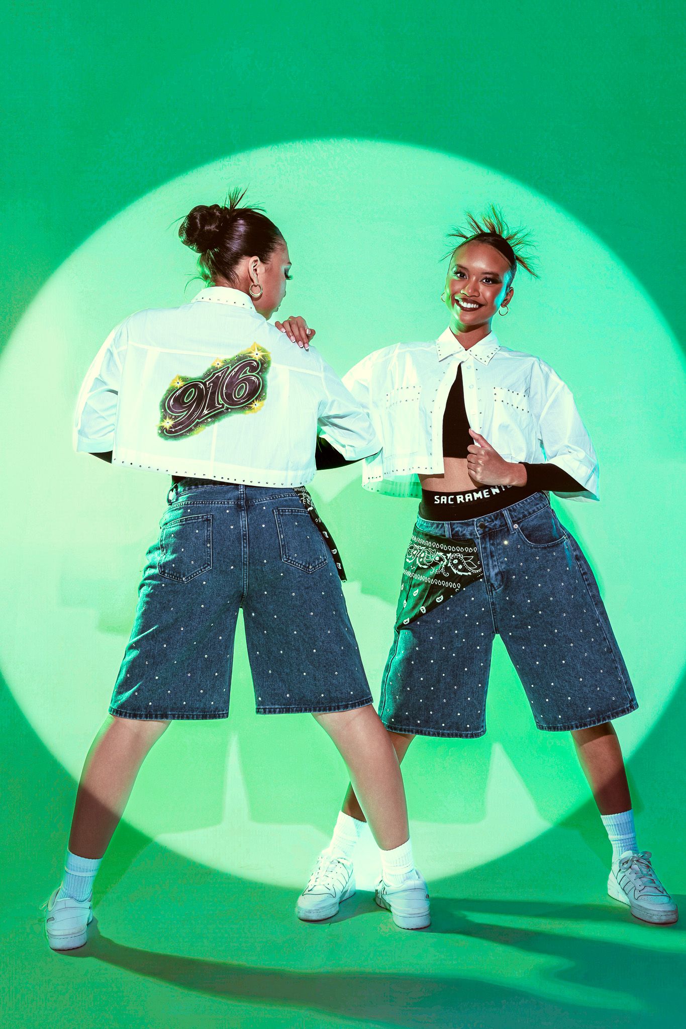 Two models wearing white cropped jackets with 916 patches and denim shorts pose against a green background.
