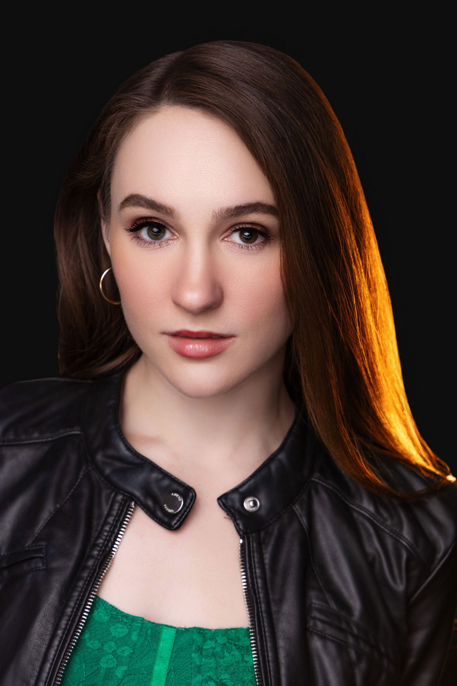 Portrait against dark background with dramatic side lighting and emerald green top under black leather jacket.