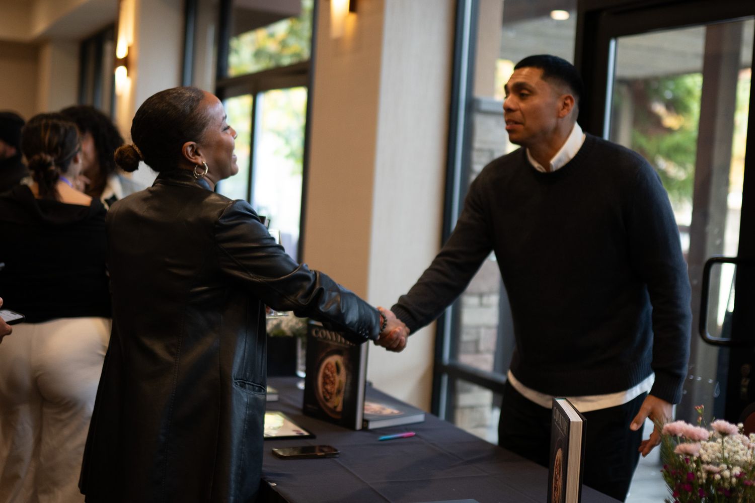 Two attendees shake hands during a social event in a modern venue with natural lighting.