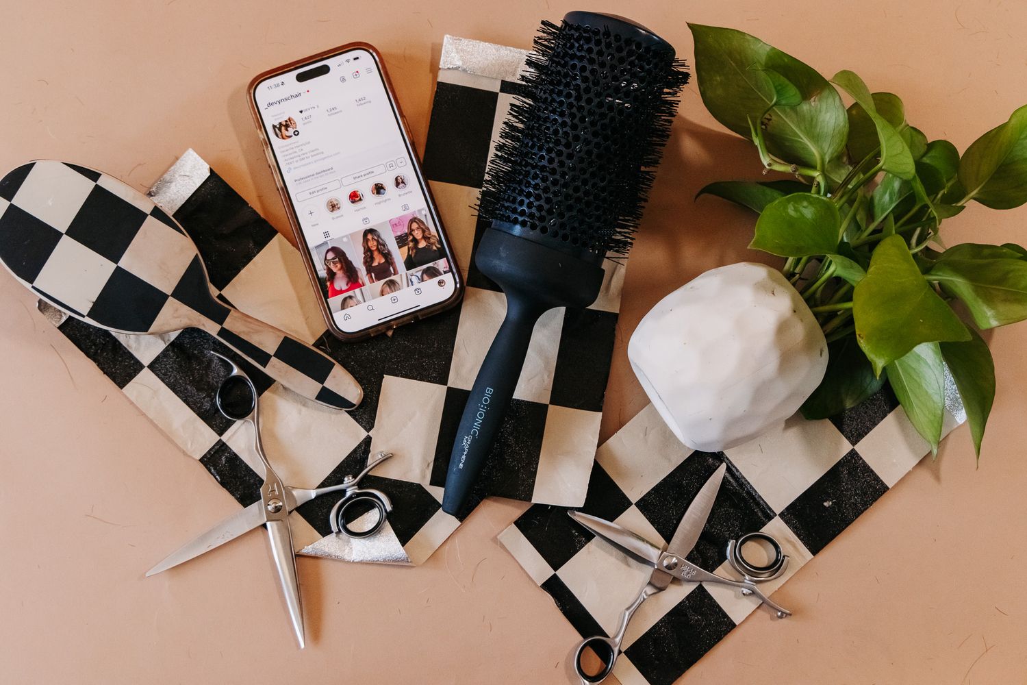 Hair styling tools and accessories including brushes, a phone, and plant arranged on a checkered black and white background.