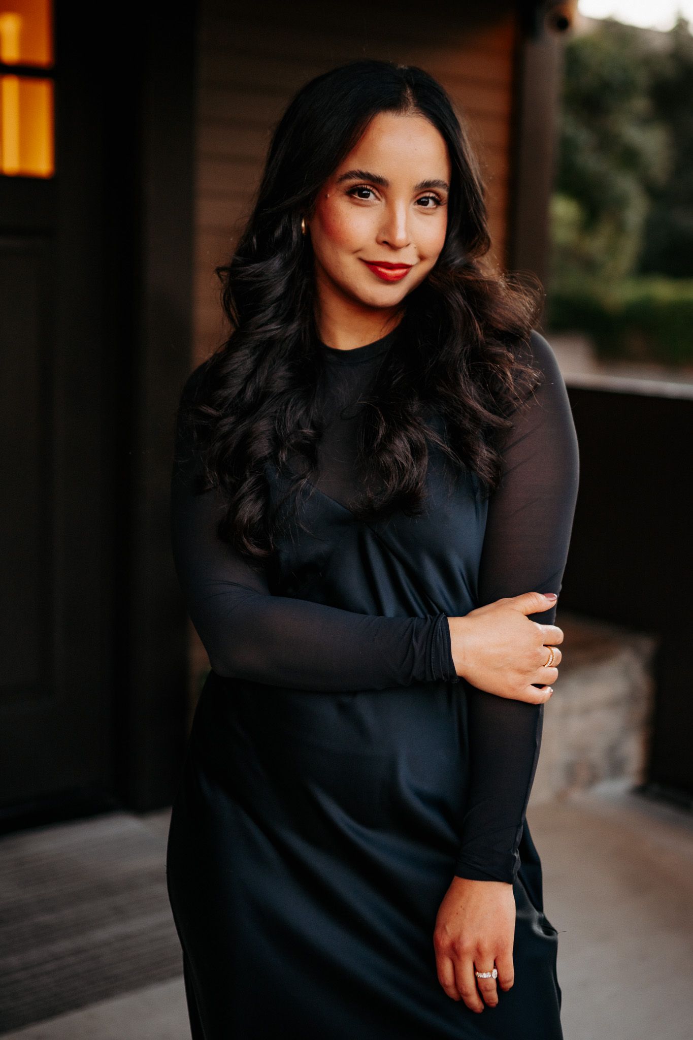 Woman in elegant black dress and sheer sleeves poses confidently on patio at dusk with warm lighting.