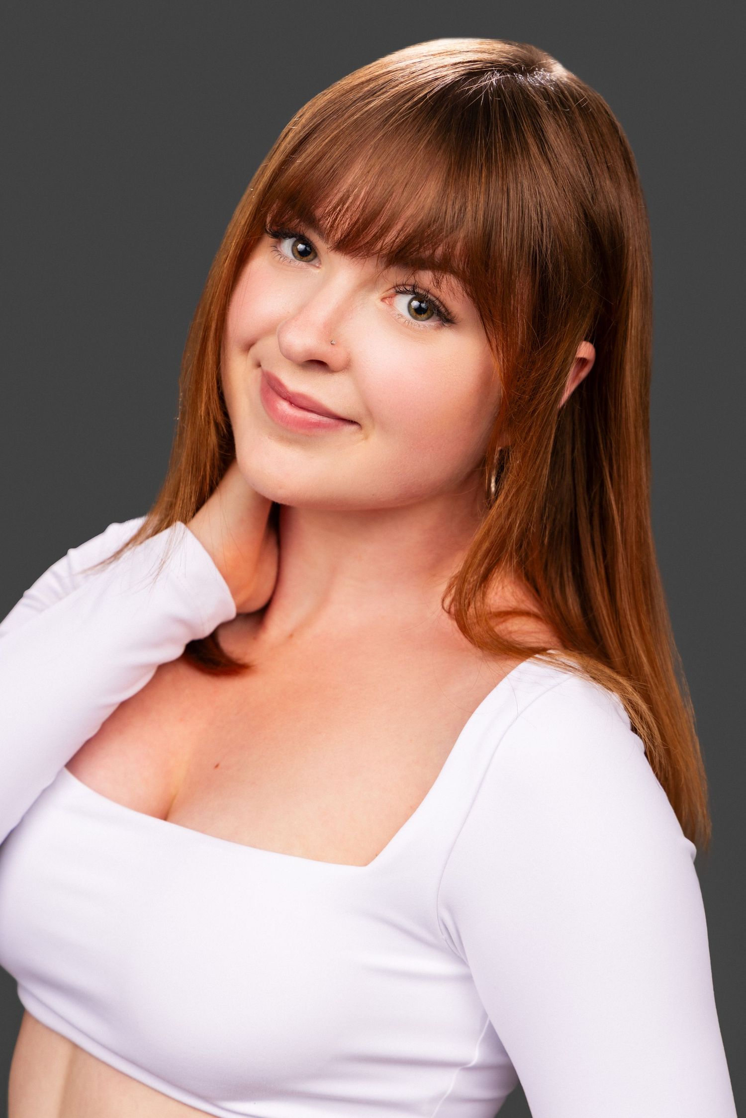 Studio portrait with straight brown hair, bangs, and white long-sleeve top against dark background.