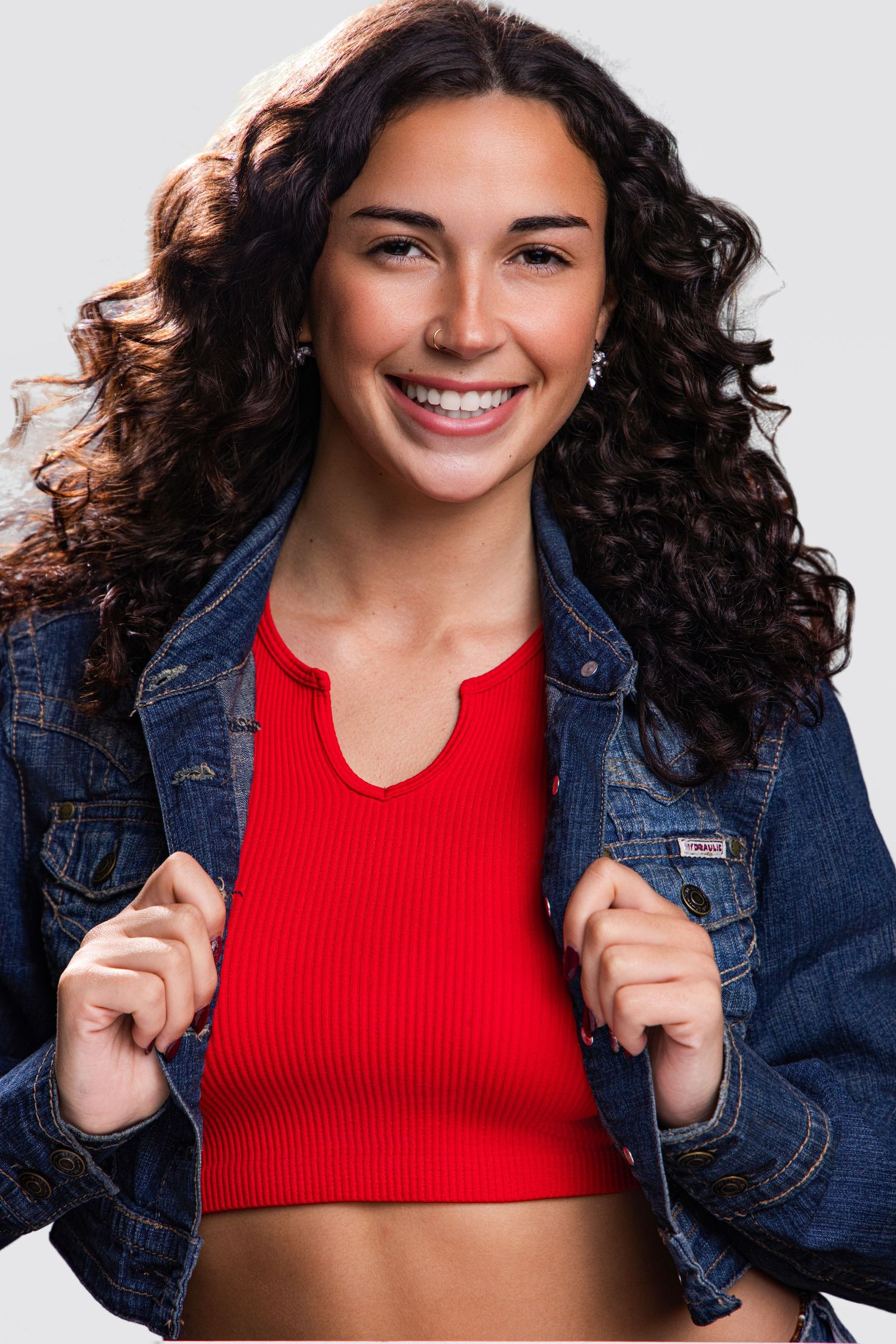 Smiling young woman with curly hair wearing a red crop top and denim jacket against a gray background.