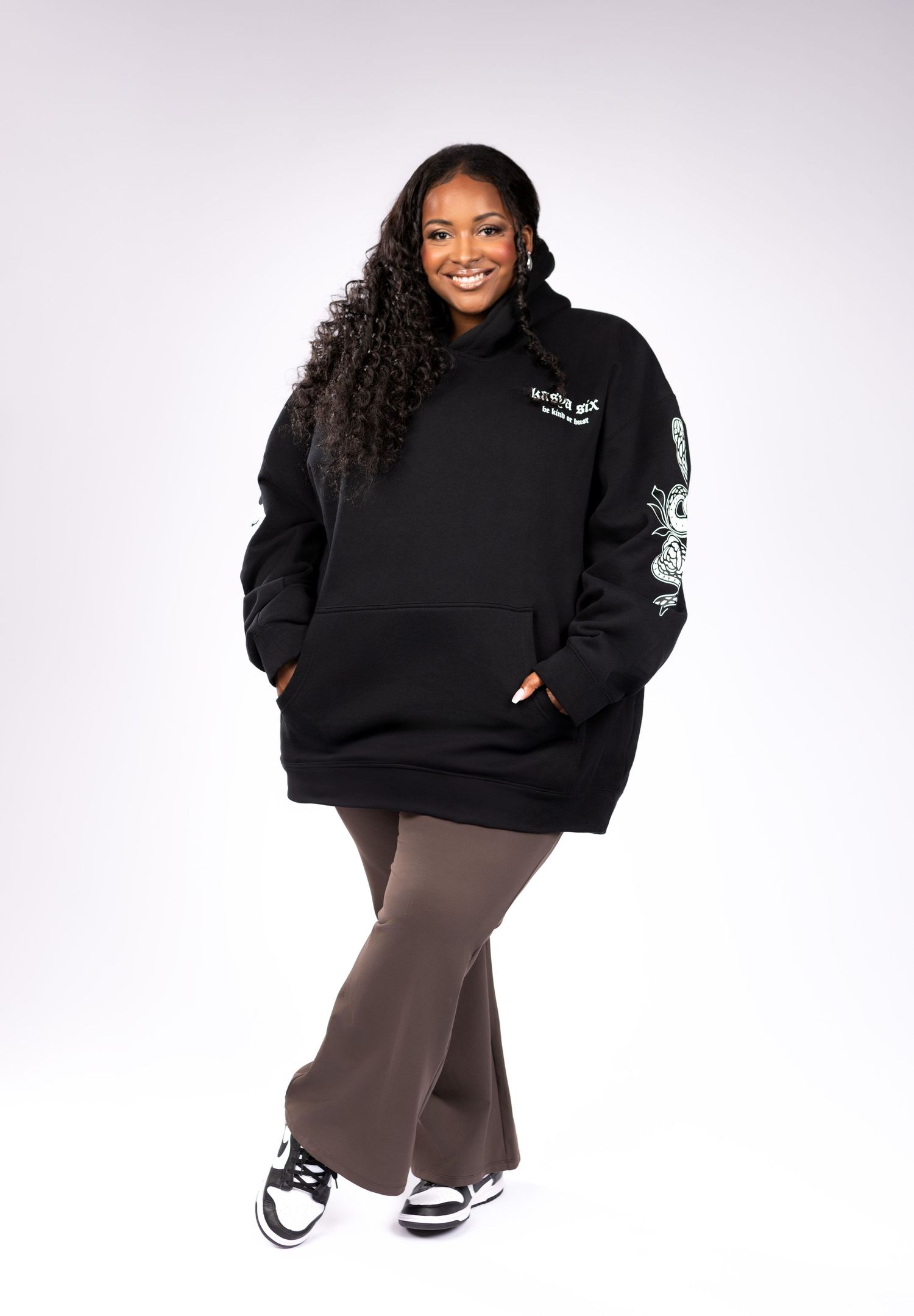 Woman smiling in oversized black dragon graphic hoodie, brown flare pants, and white sneakers.
