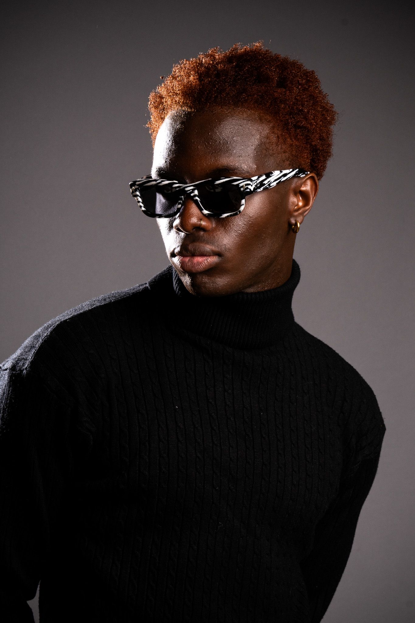 Young Black man with dyed red curly hair wearing black sunglasses and turtleneck sweater poses against gray background.