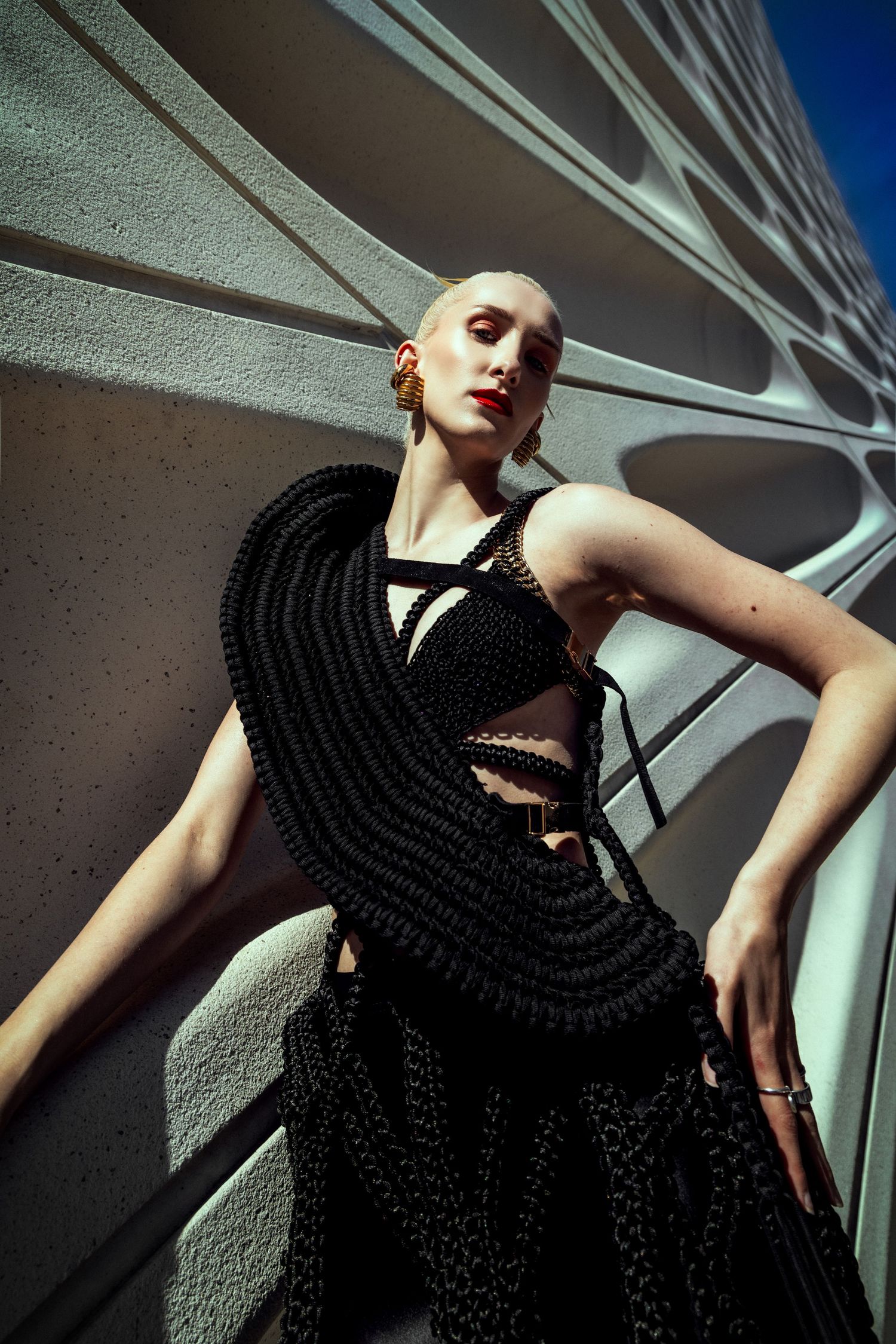 Model in avant-garde black woven sculptural dress poses against modern architectural white wall in dramatic fashion editorial.