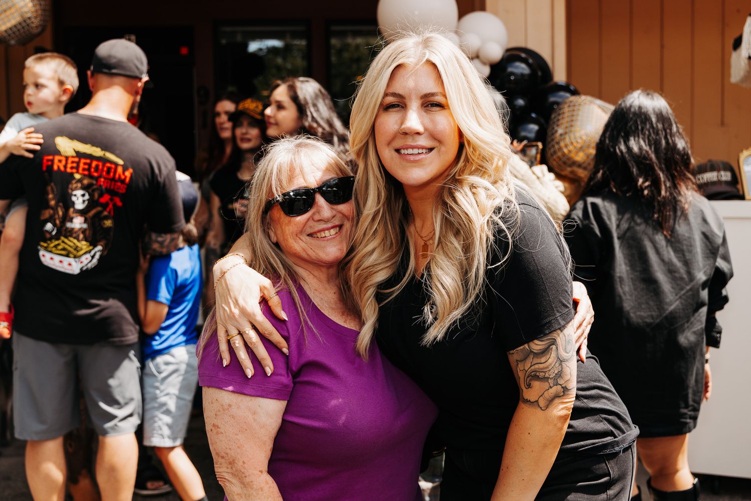 Two women smiling and hugging outdoors at a casual backyard gathering with balloons in background.