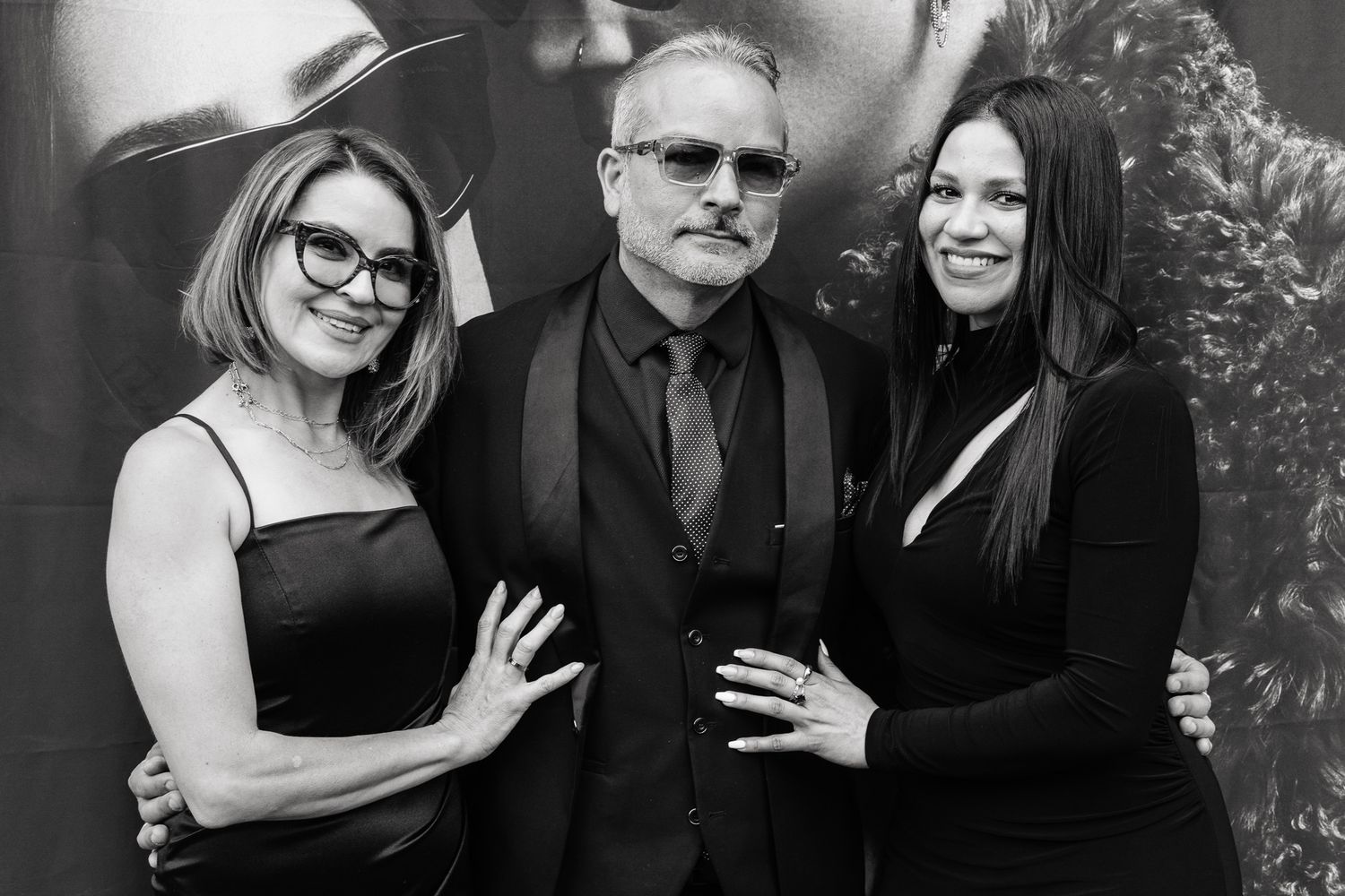 A man in a black suit poses with two women in black dresses at a formal event in a black and white photo.