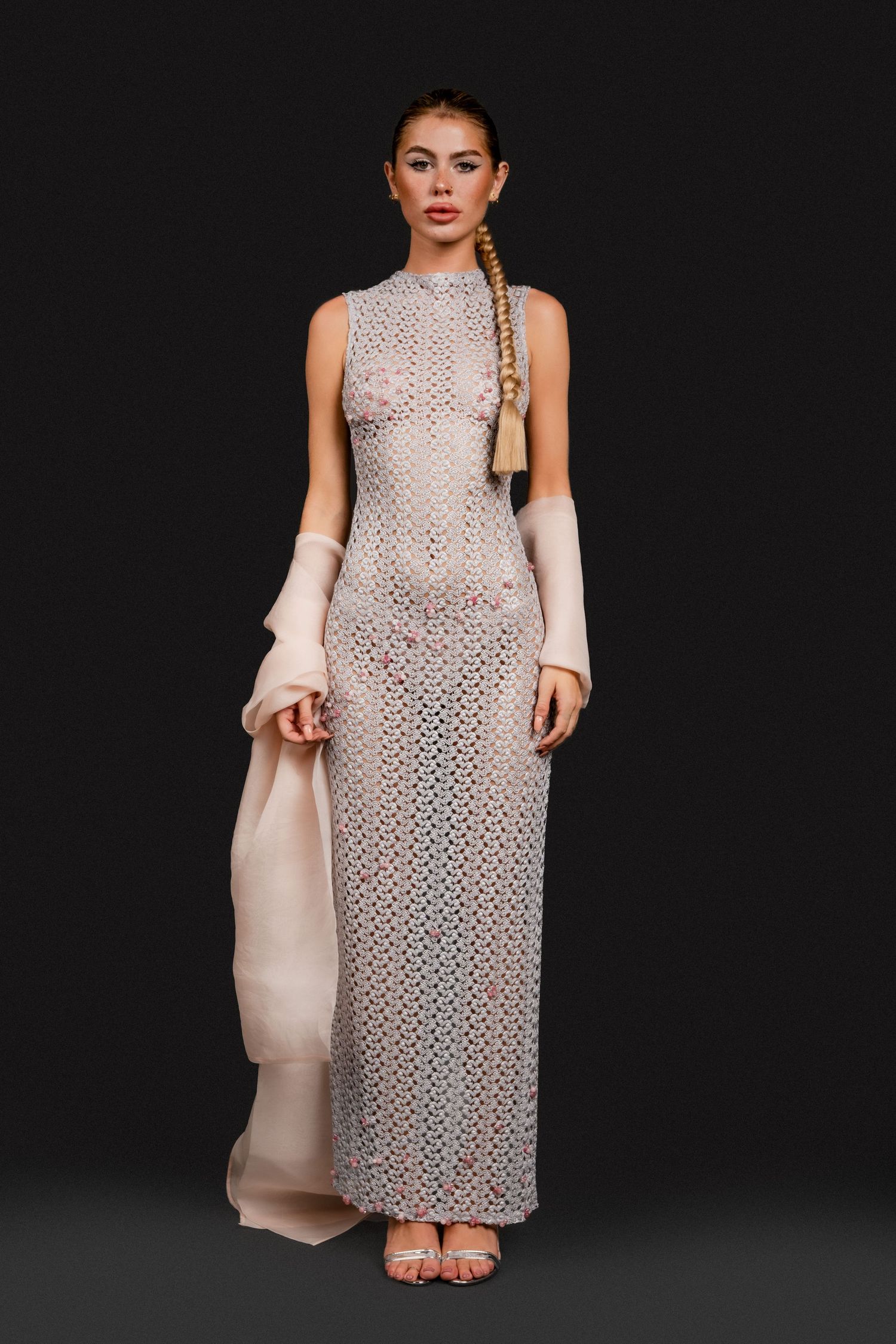 Model in a sleeveless silver crochet maxi dress with pink accents, long braided hair, and beige arm wraps.